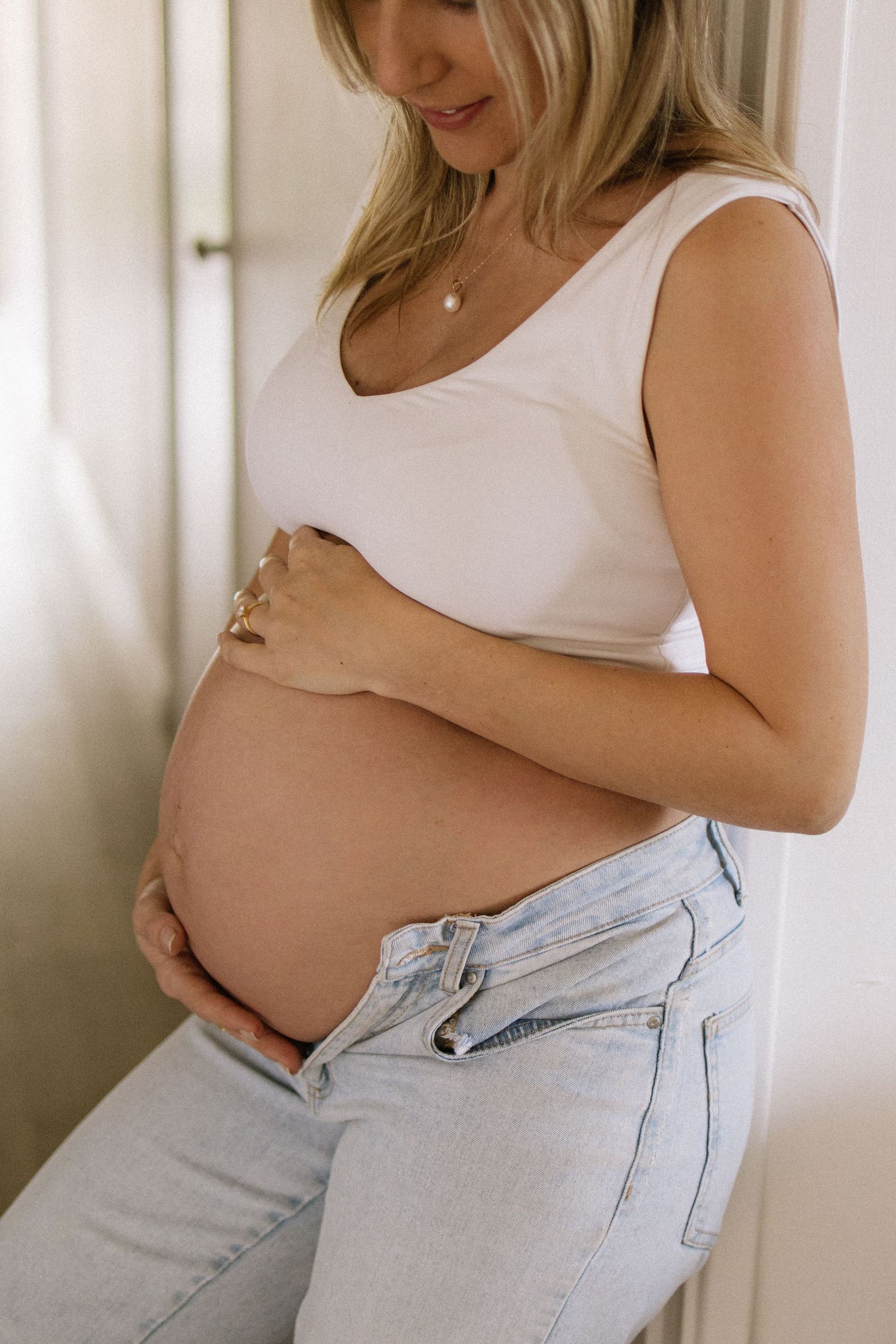 Pregnant blonde woman in white tank top and jeans gently cradles her bare baby bump.