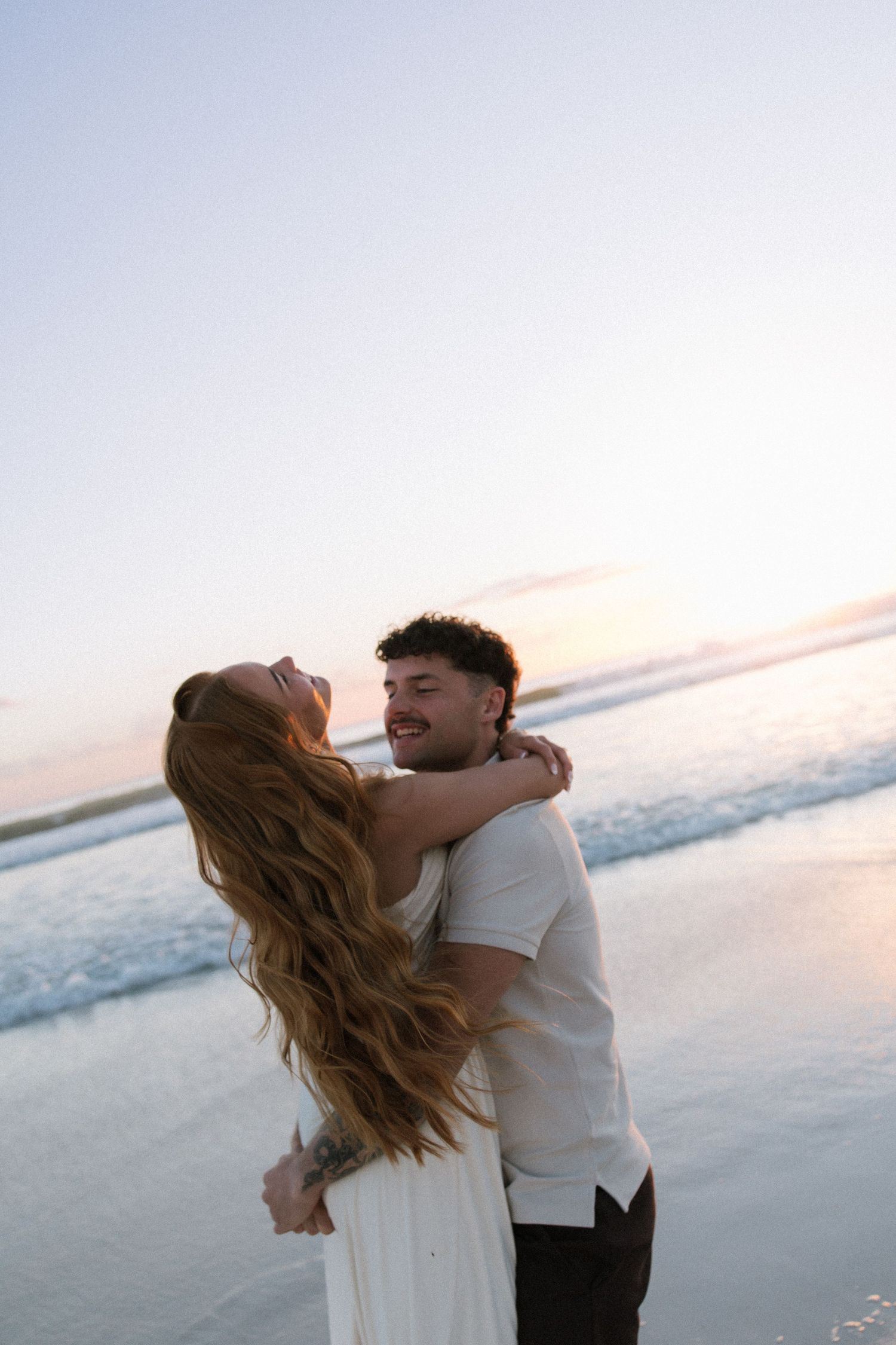 Smiling couple embracing on beach at sunset, woman in white dress with long hair.