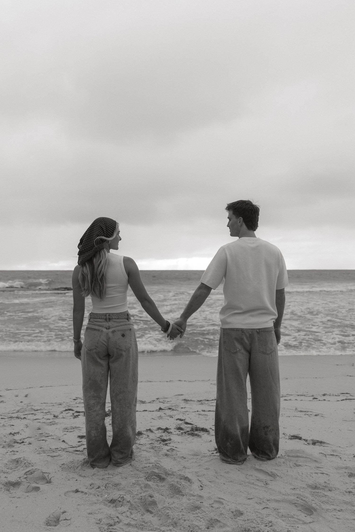 Black and white rear view of a couple holding hands while standing at the ocean's edge.