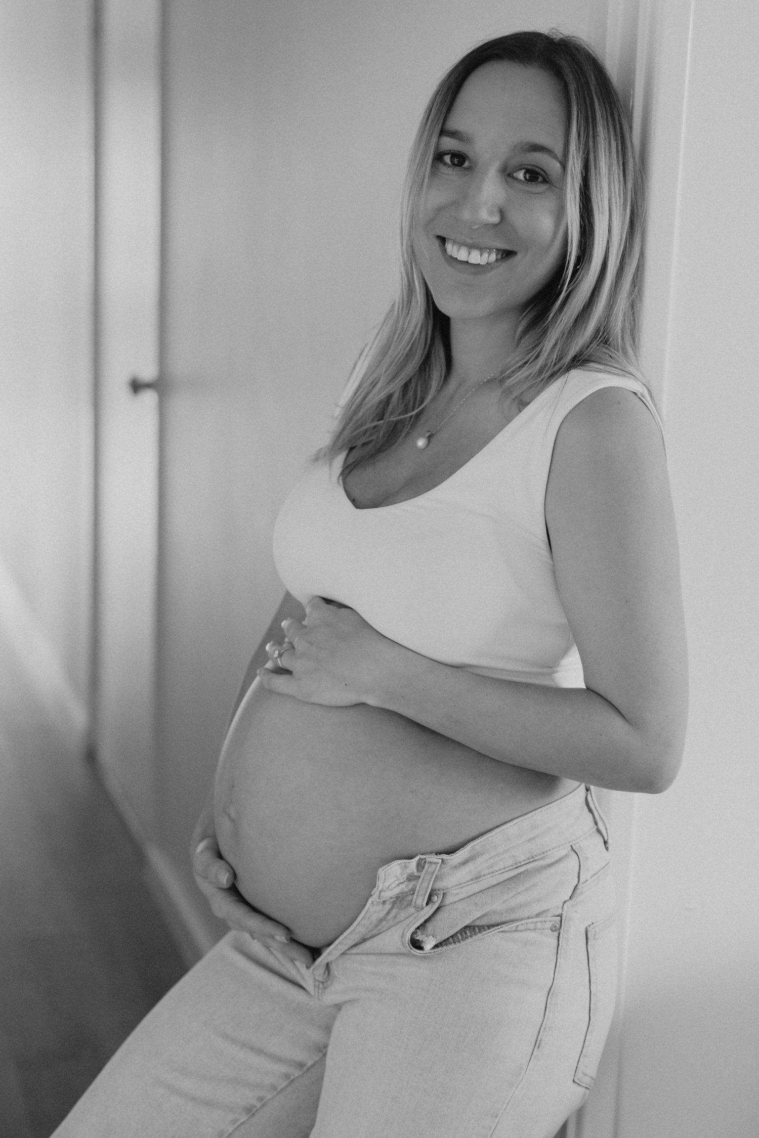 Smiling pregnant blonde woman in white tank top holds baby bump in black and white portrait.