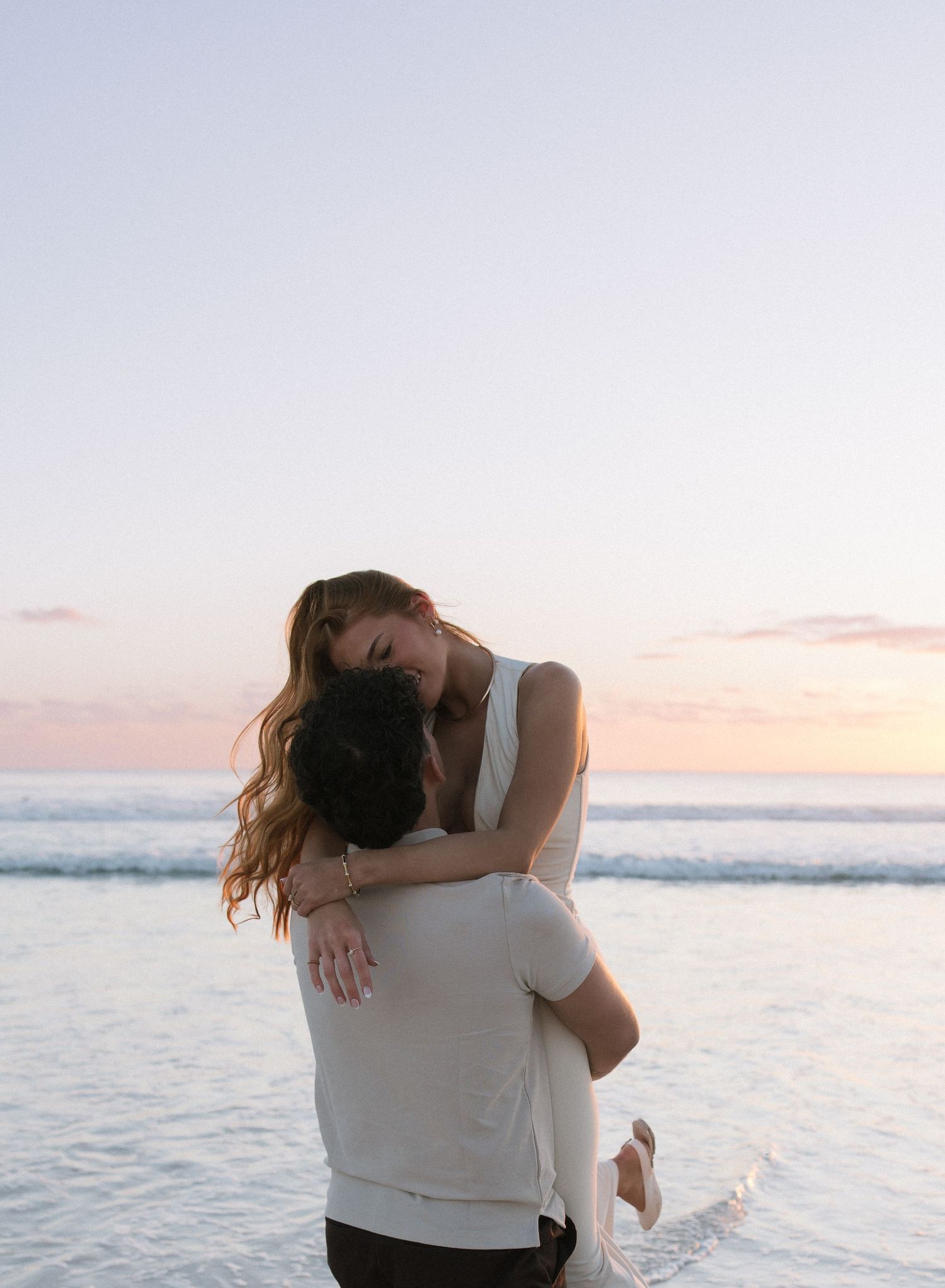 Groom lifting bride in white dress during romantic beach sunset embrace.