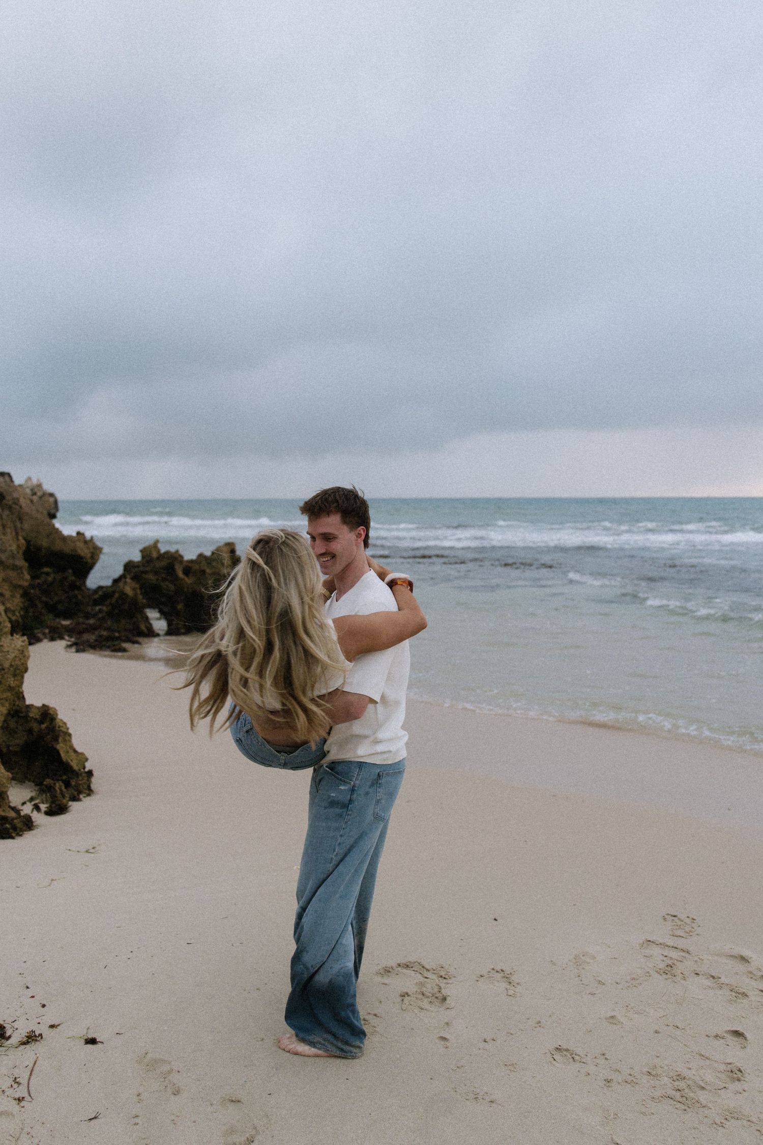 Happy couple embracing on a sandy beach with rocky coastline and ocean waves behind them.