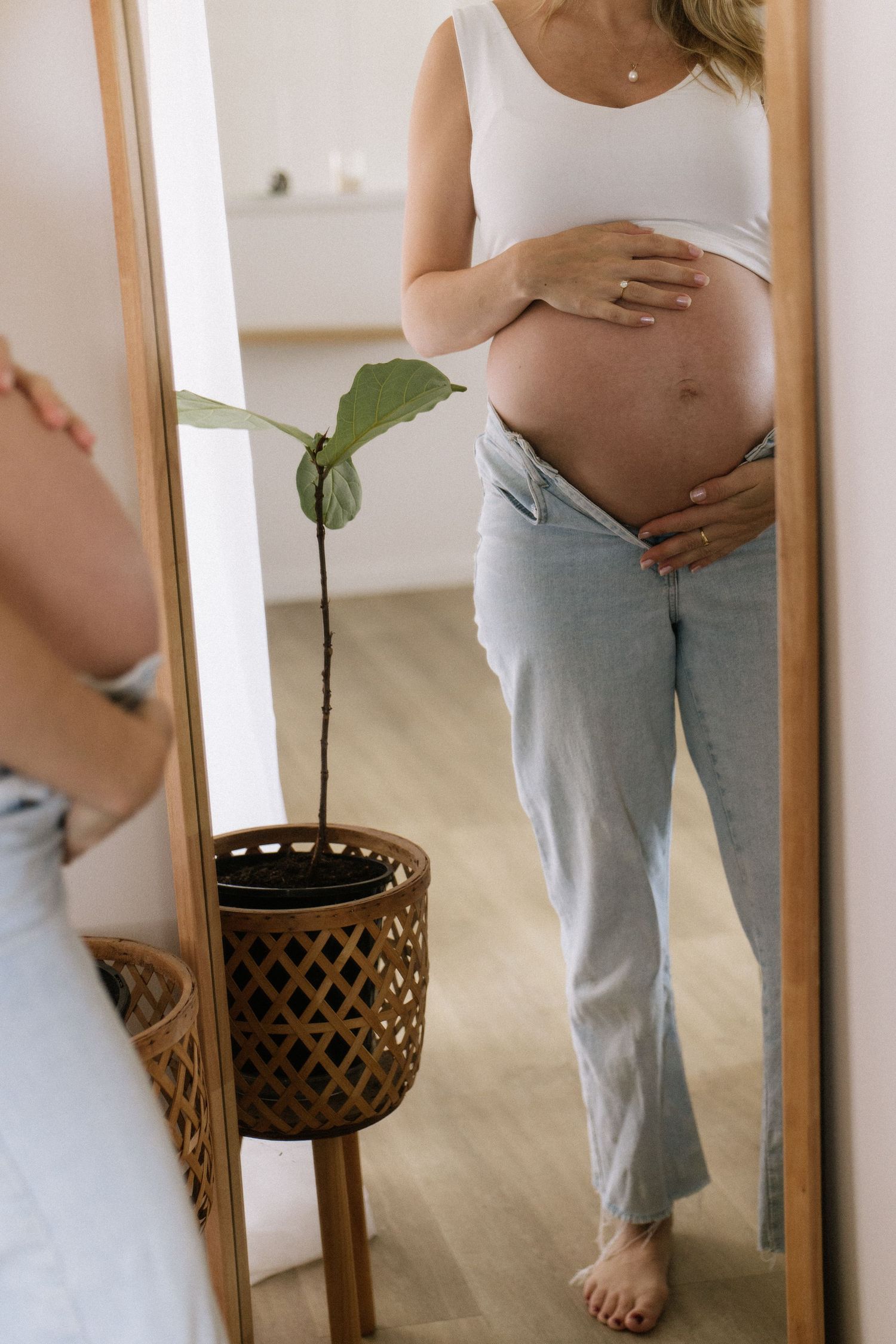 Pregnant woman in a white tank top and jeans examining her baby bump in a full-length wooden mirror.