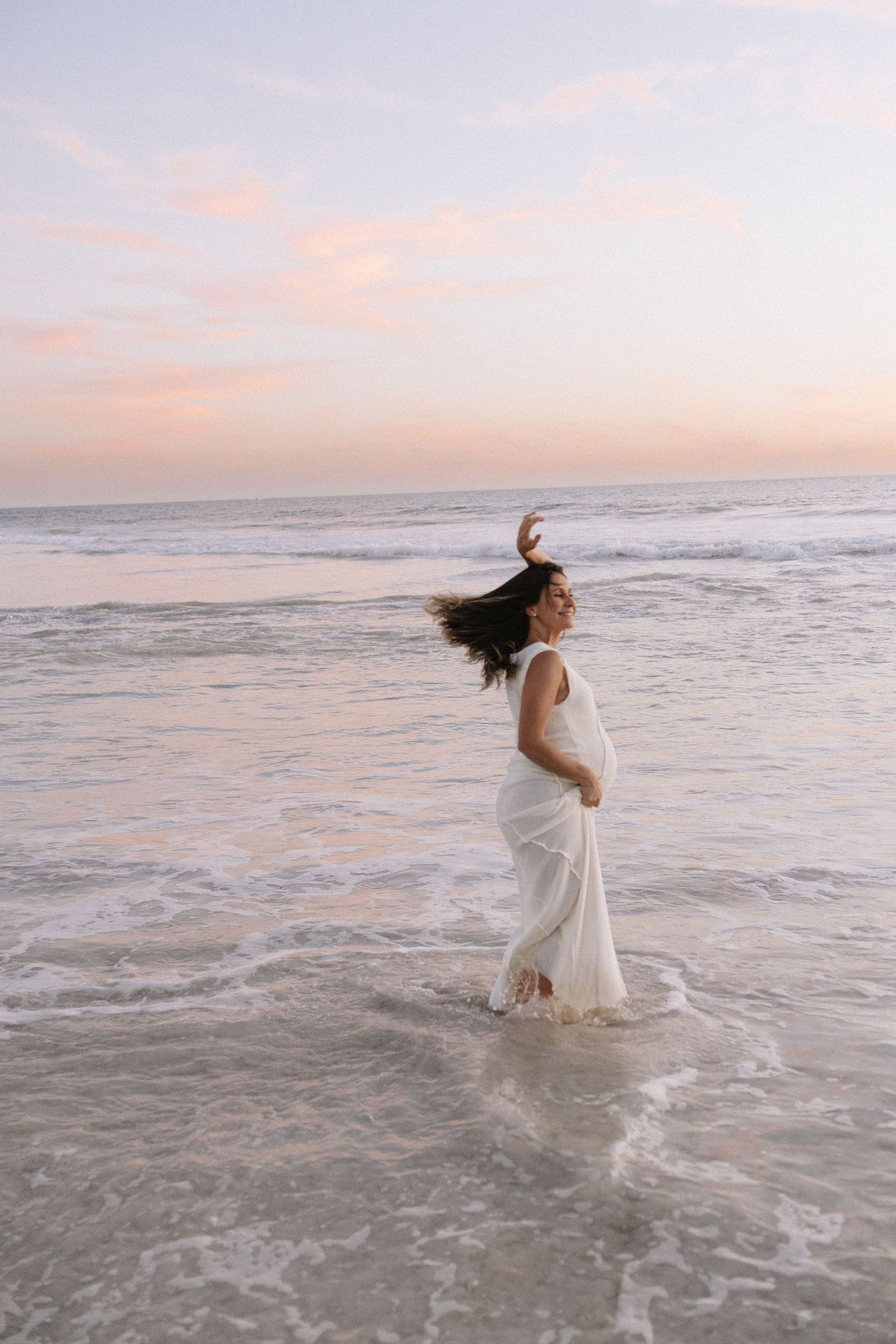 Pregnant woman in white dress standing in ocean waves at sunset.