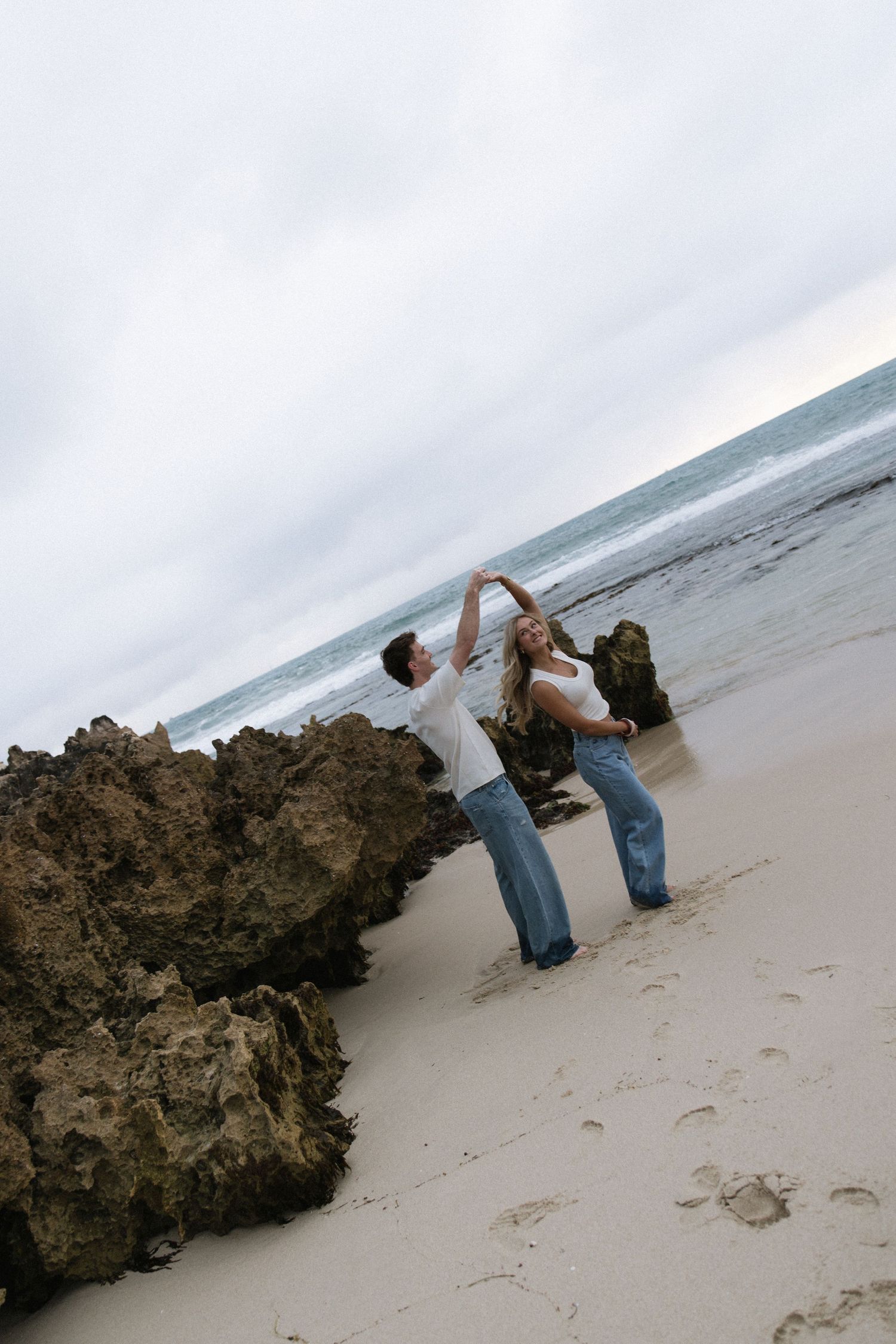 Couple dancing together on a sandy beach near rocky cliffs with overcast sky and ocean in background.
