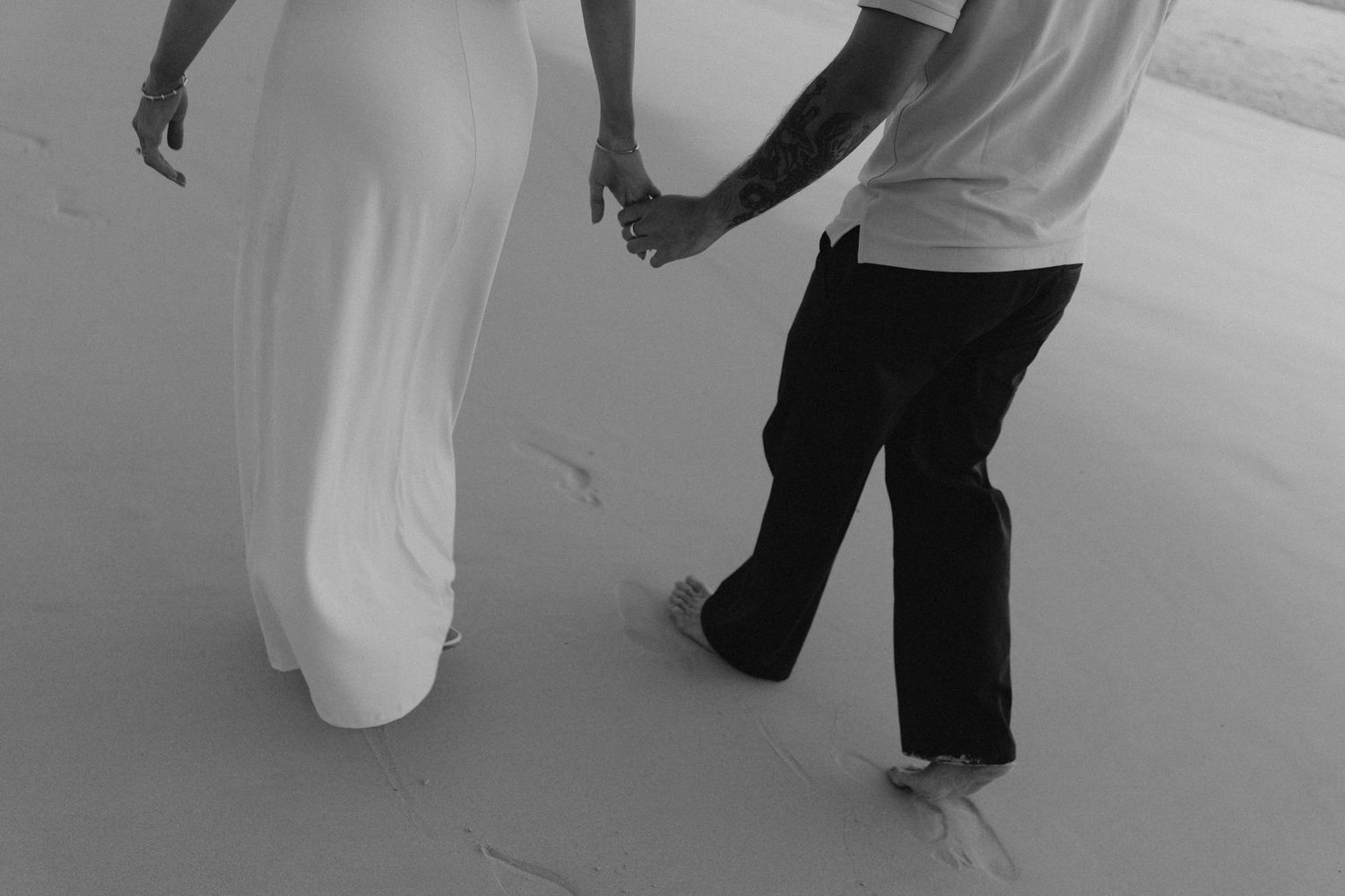 Black and white aerial view of couple holding hands while walking barefoot on sandy beach.