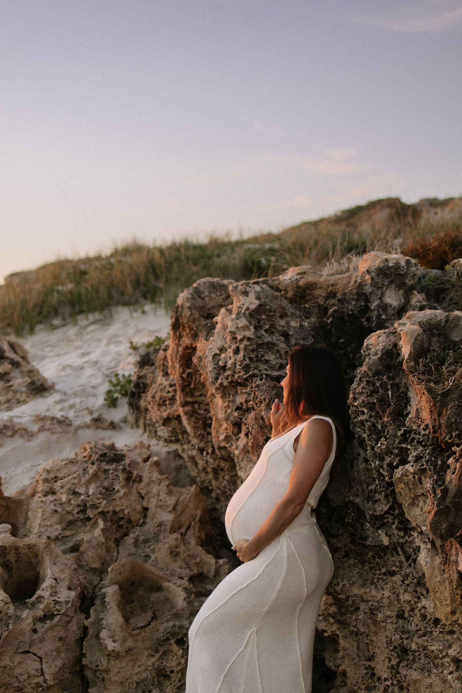 Pregnant woman in white dress leaning against coastal rocks at sunset beach.