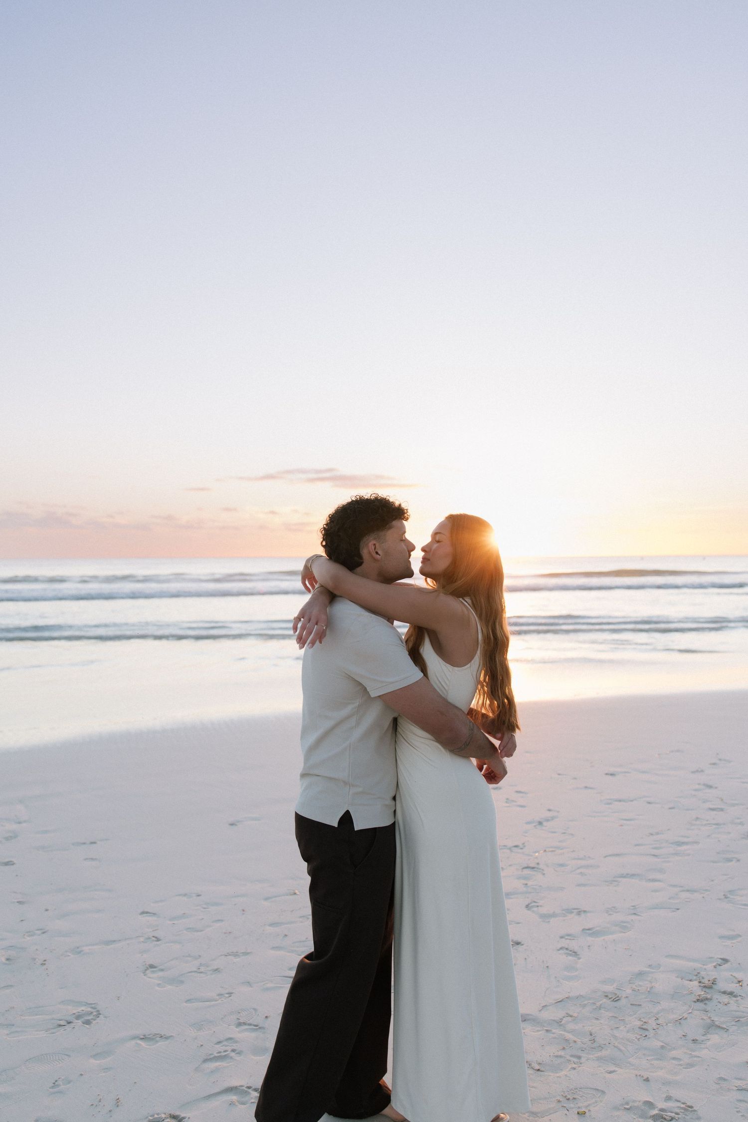 Couple embracing and almost kissing on a sandy beach at sunset, woman in elegant white dress.