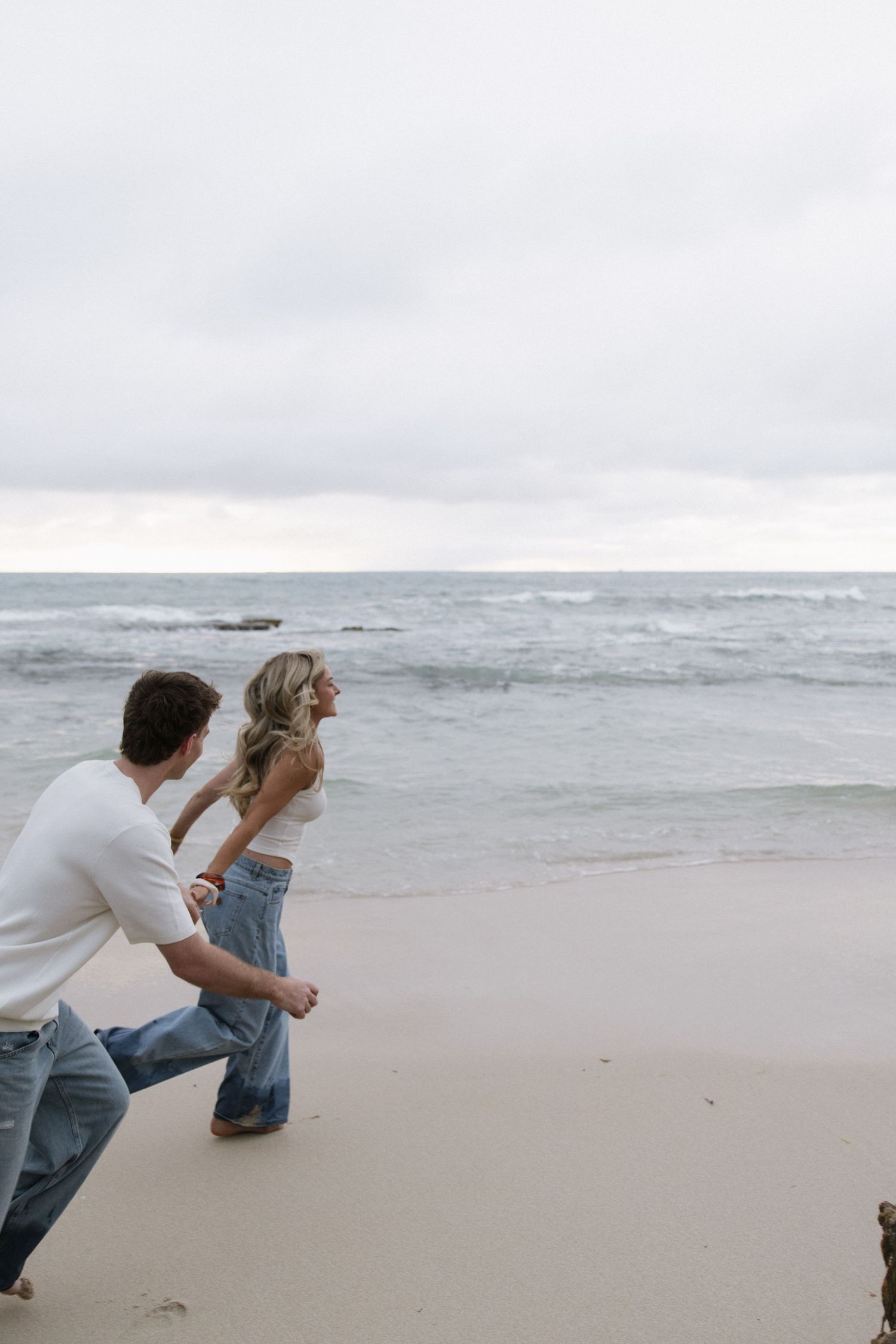 Couple running playfully on a sandy beach with ocean waves in the background on a cloudy day.
