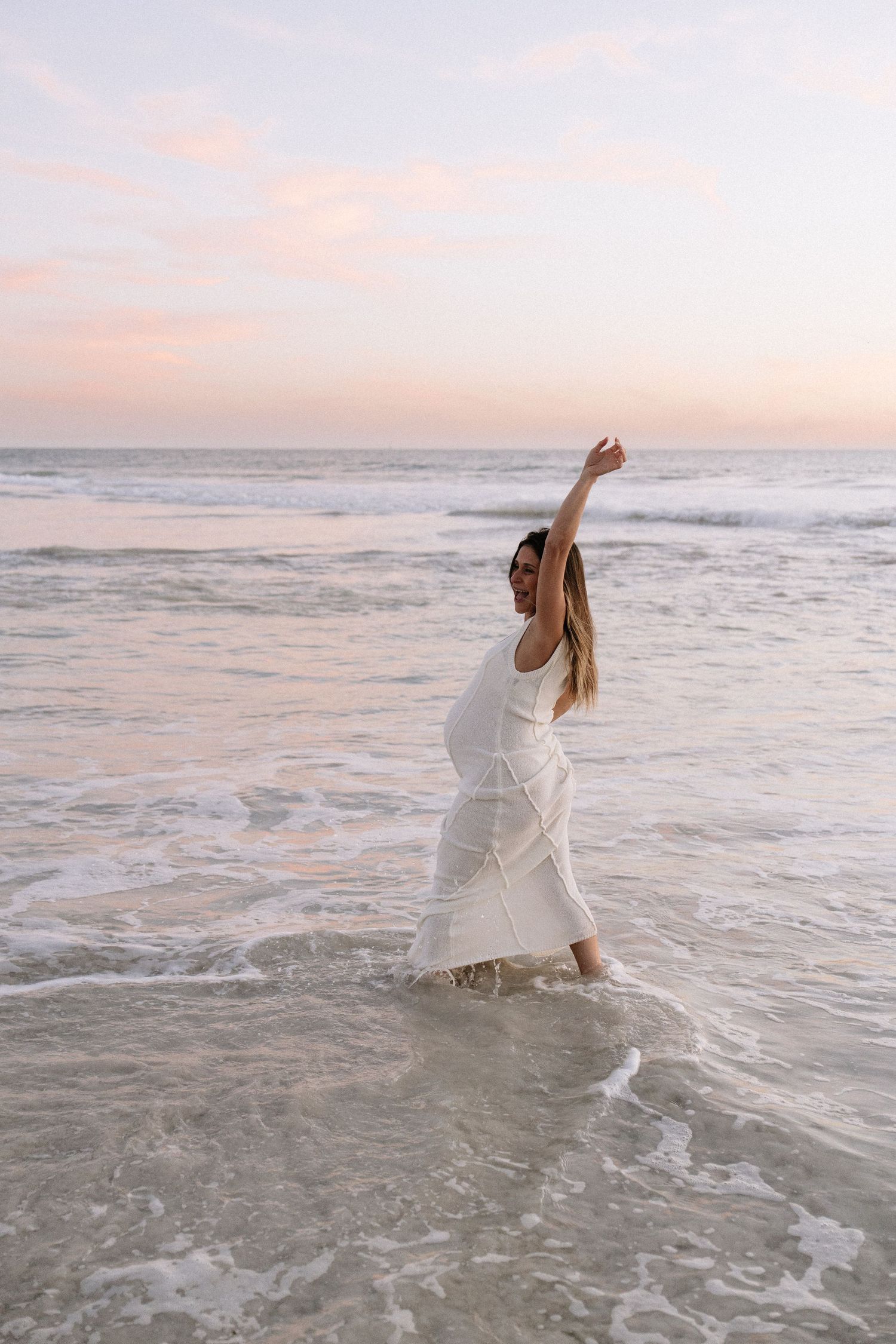 Pregnant woman in white dress raising arm while standing in ocean waves.