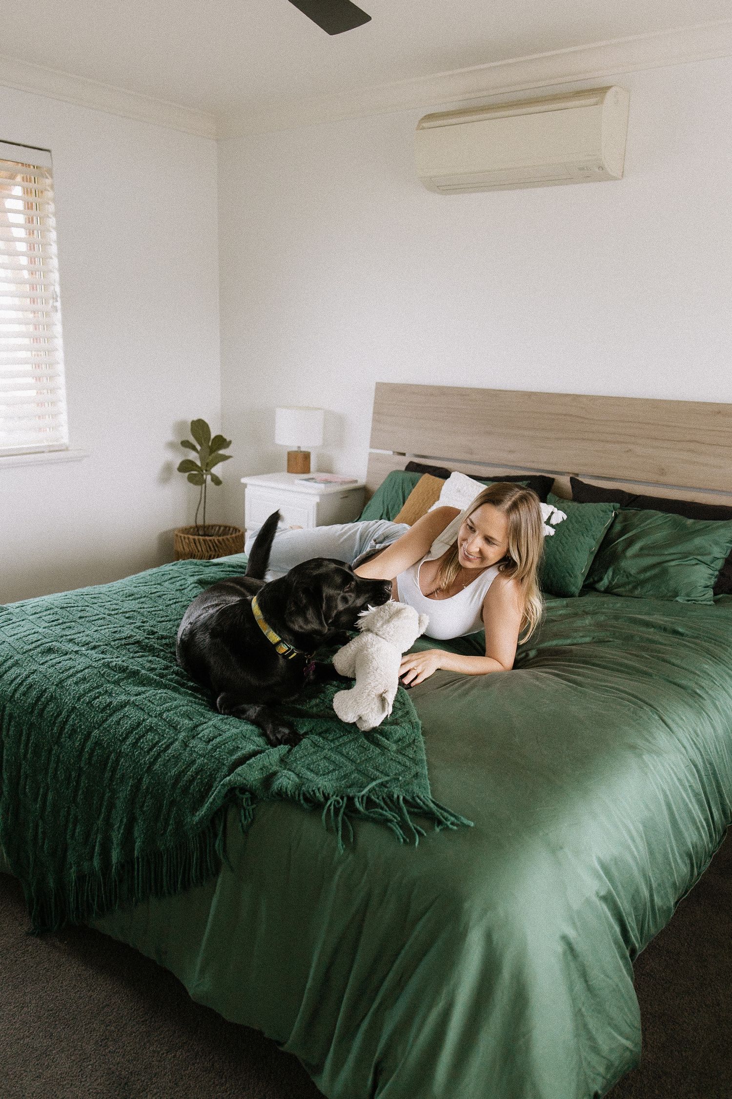 Smiling pregnant woman in white top lies on green bedding playing with black Labrador dog.