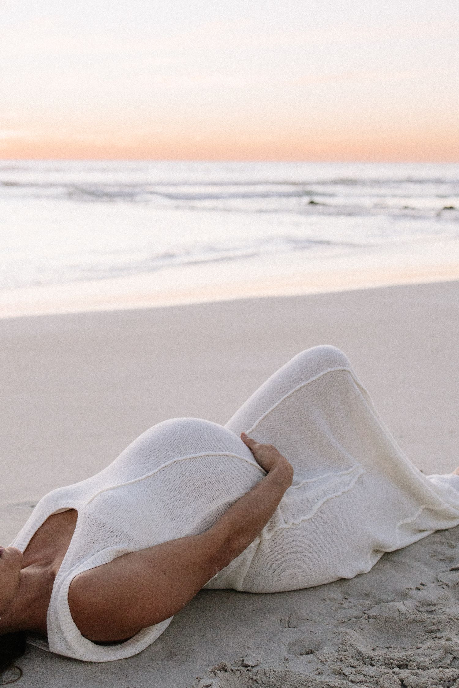 Pregnant woman in white dress lying on sandy beach at sunset by ocean waves.
