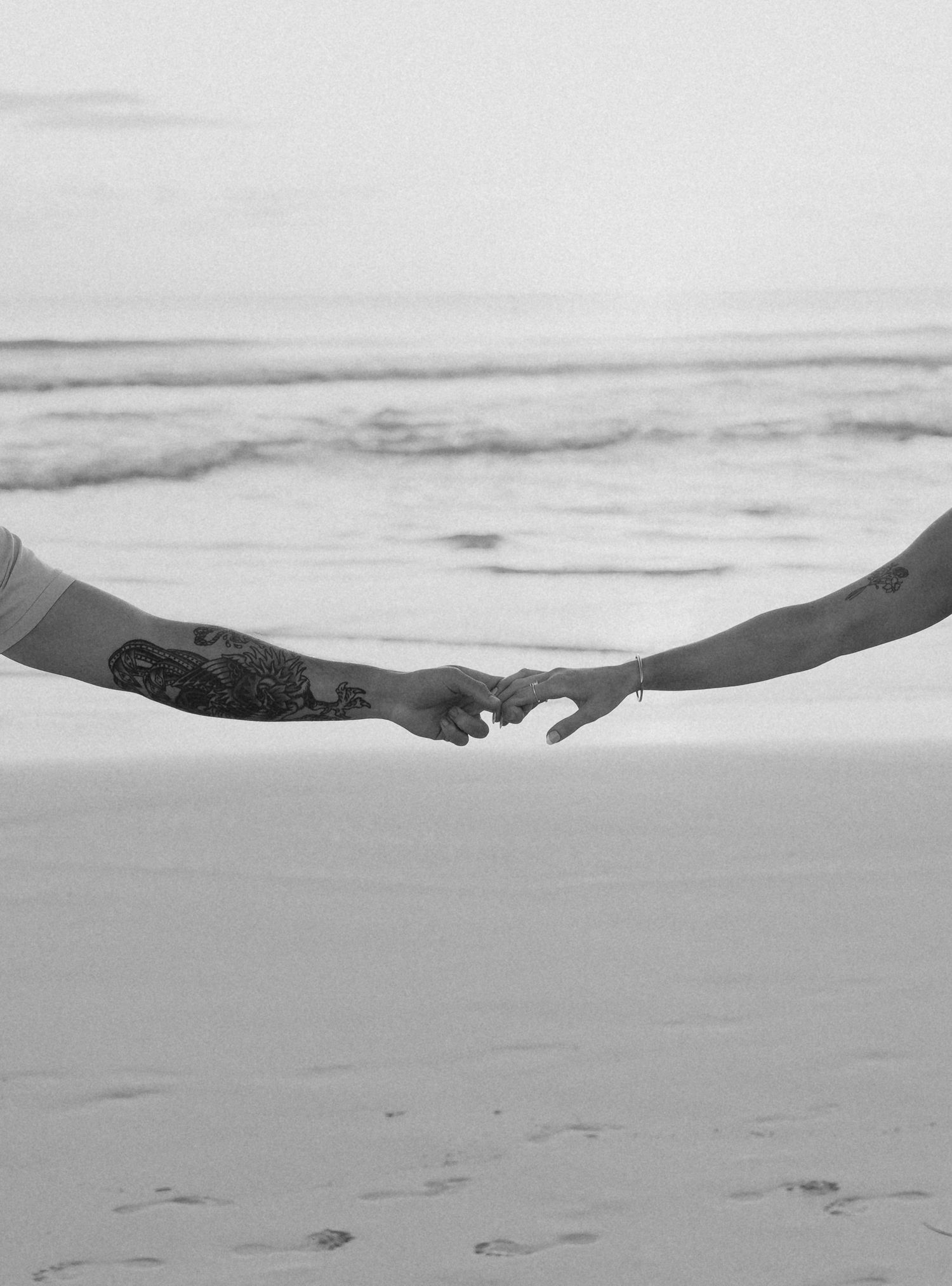 Black and white close-up of couple holding fingertips apart on beach, man with arm tattoo visible.