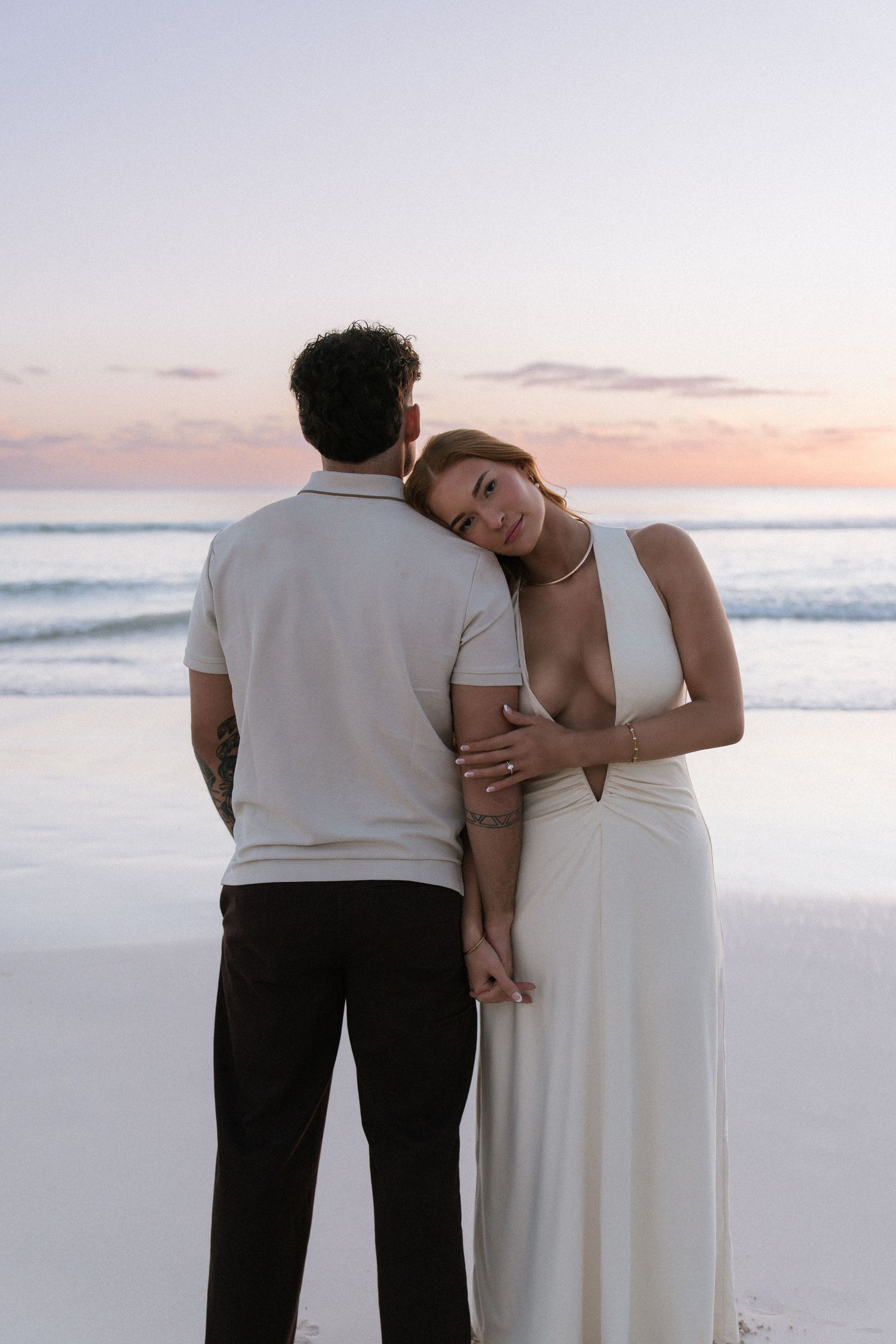 Woman in white dress leaning head on partners shoulder at beach sunset, smiling at camera.