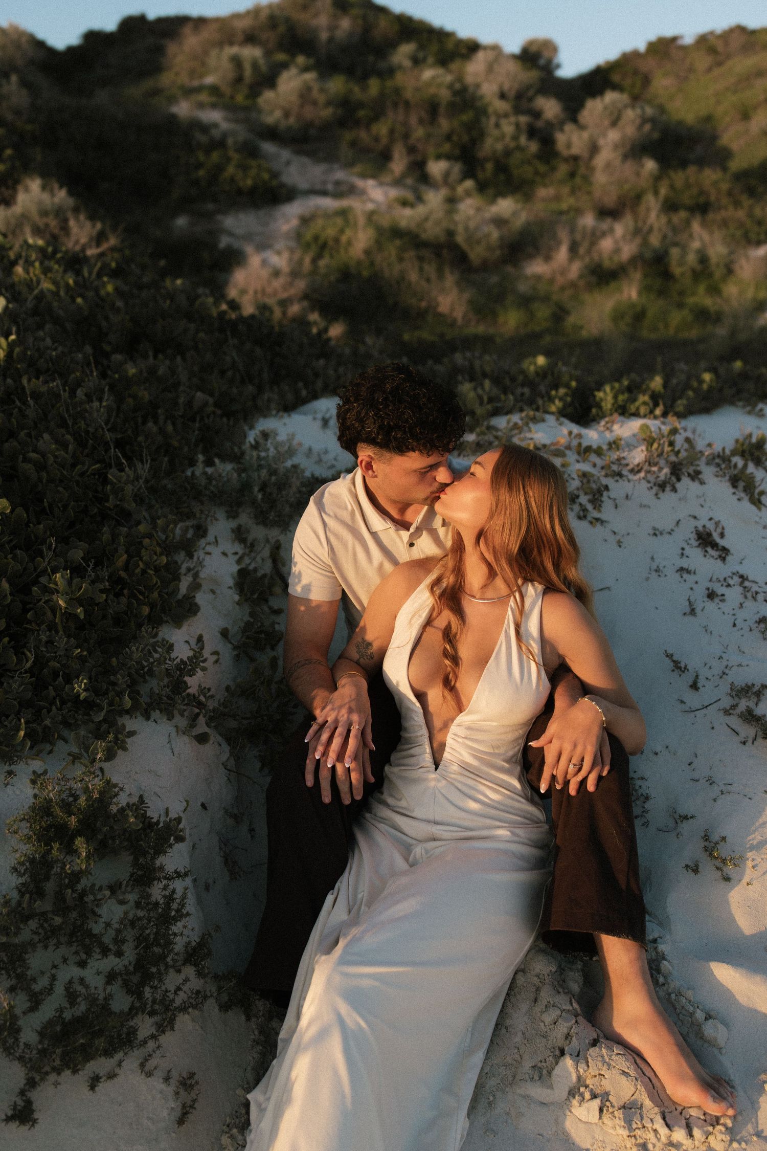 Couple sharing a romantic moment on white sand dunes surrounded by lush green coastal shrubs.