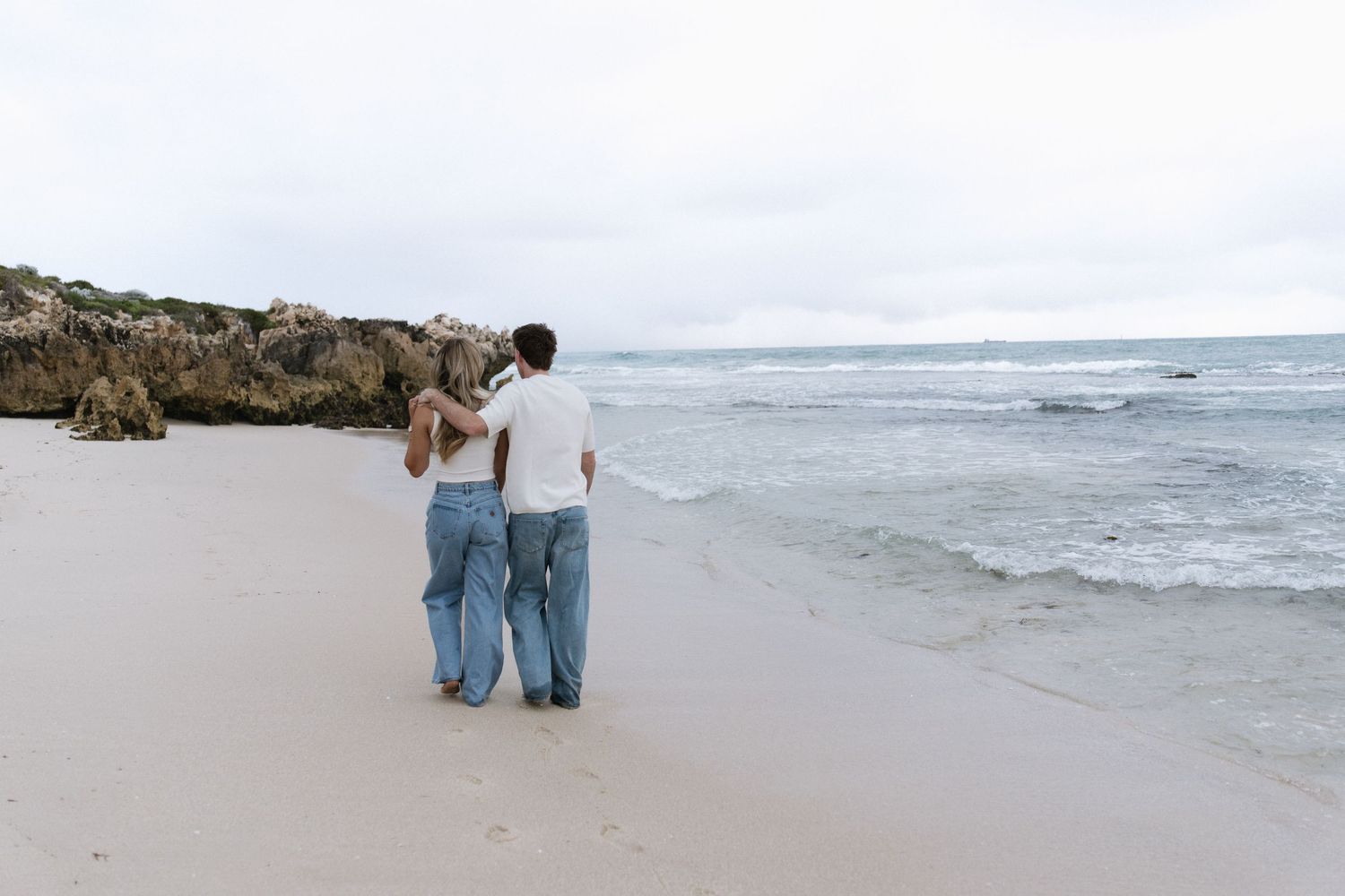 Couple walking hand in hand along a sandy beach with rocky cliffs and ocean waves in the background.