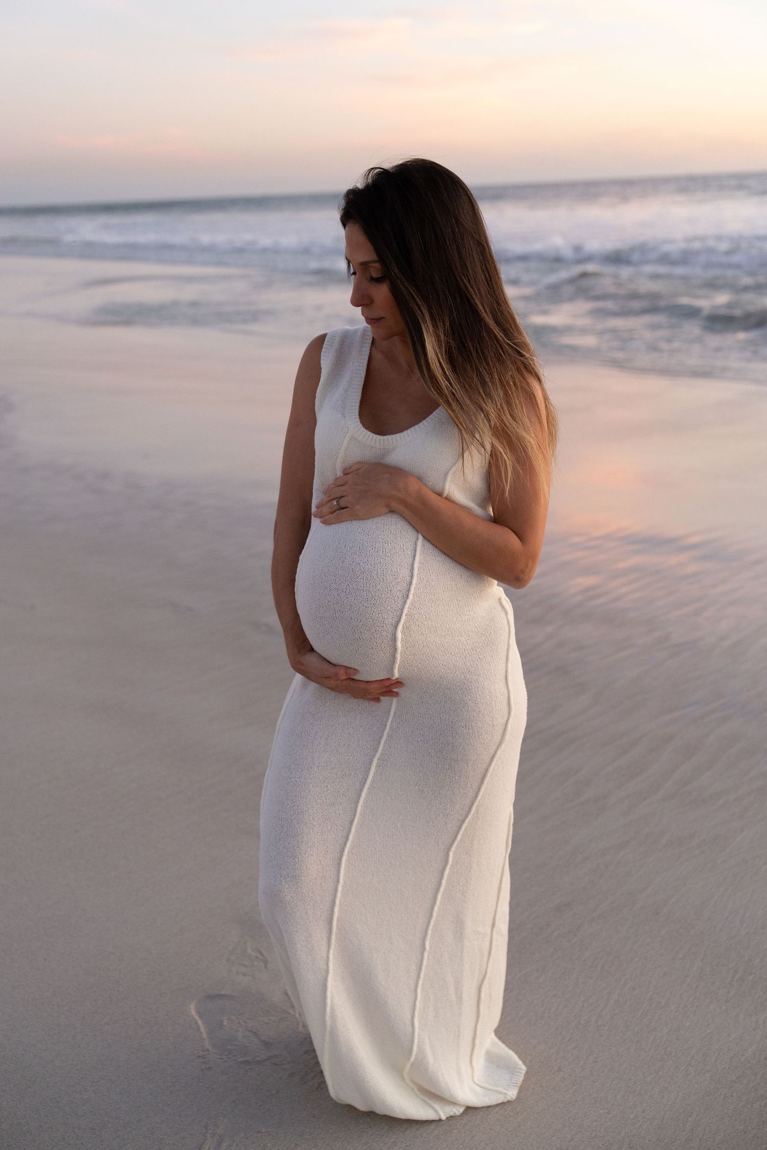 Pregnant woman in white dress holding belly on beach at sunset.