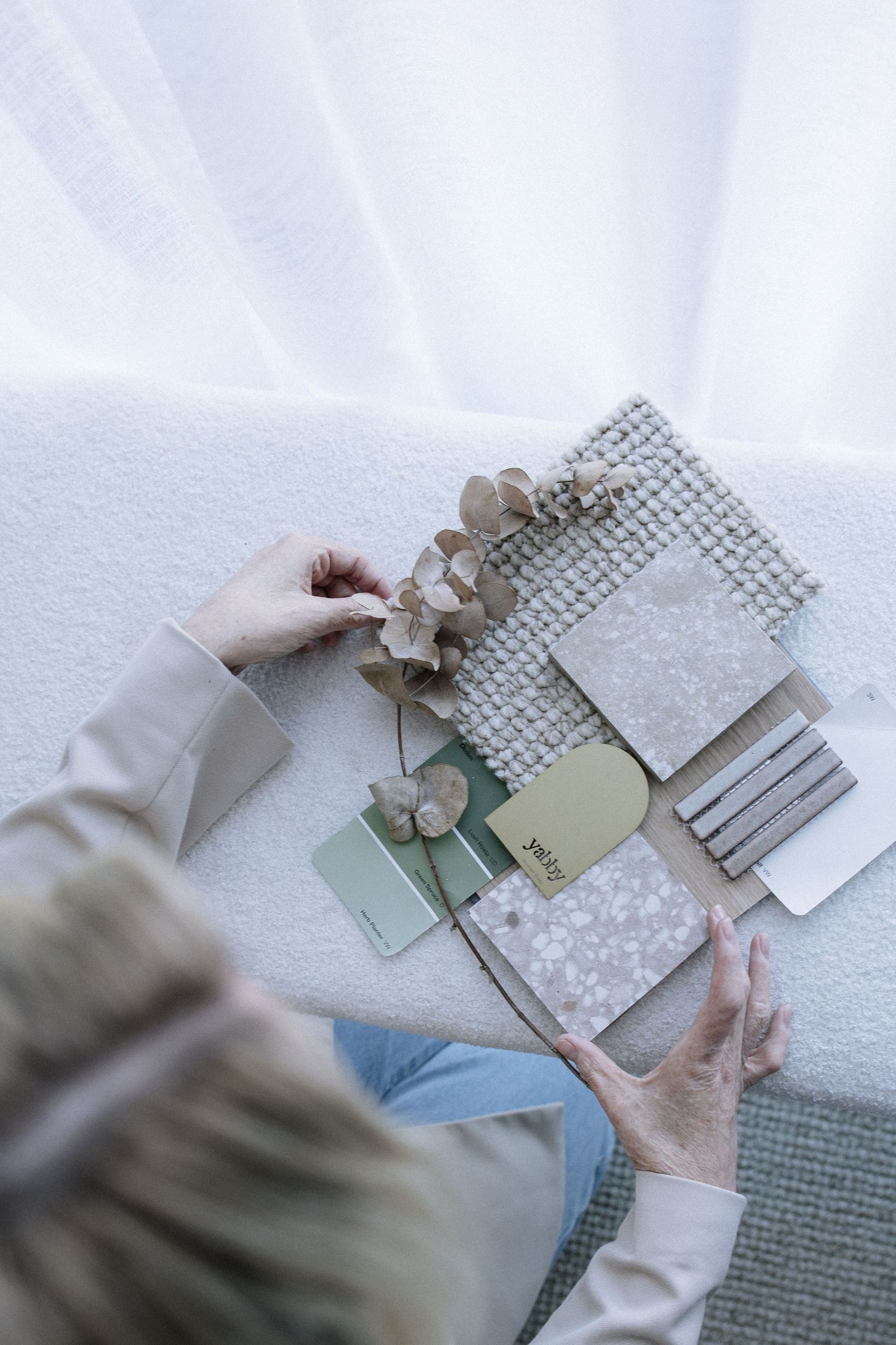 Interior designer arranging material samples, paint swatches, and dried flowers on a white textured surface.