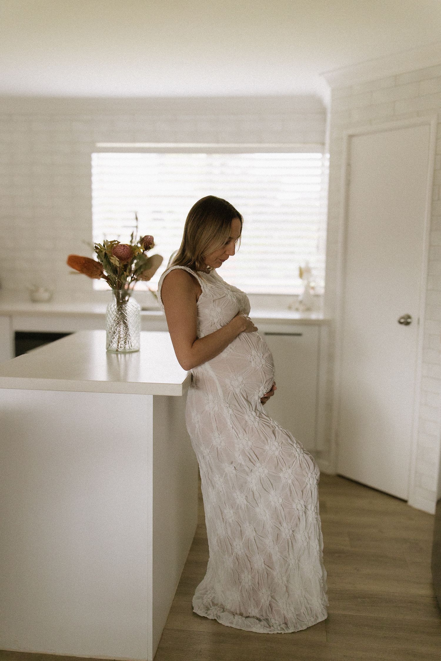 Pregnant woman in white lace dress standing by kitchen counter holding her baby bump.