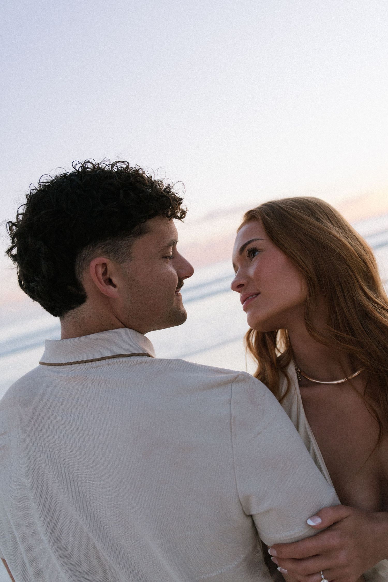 Engaged couple gazing at each other lovingly on a beach during golden hour sunset.