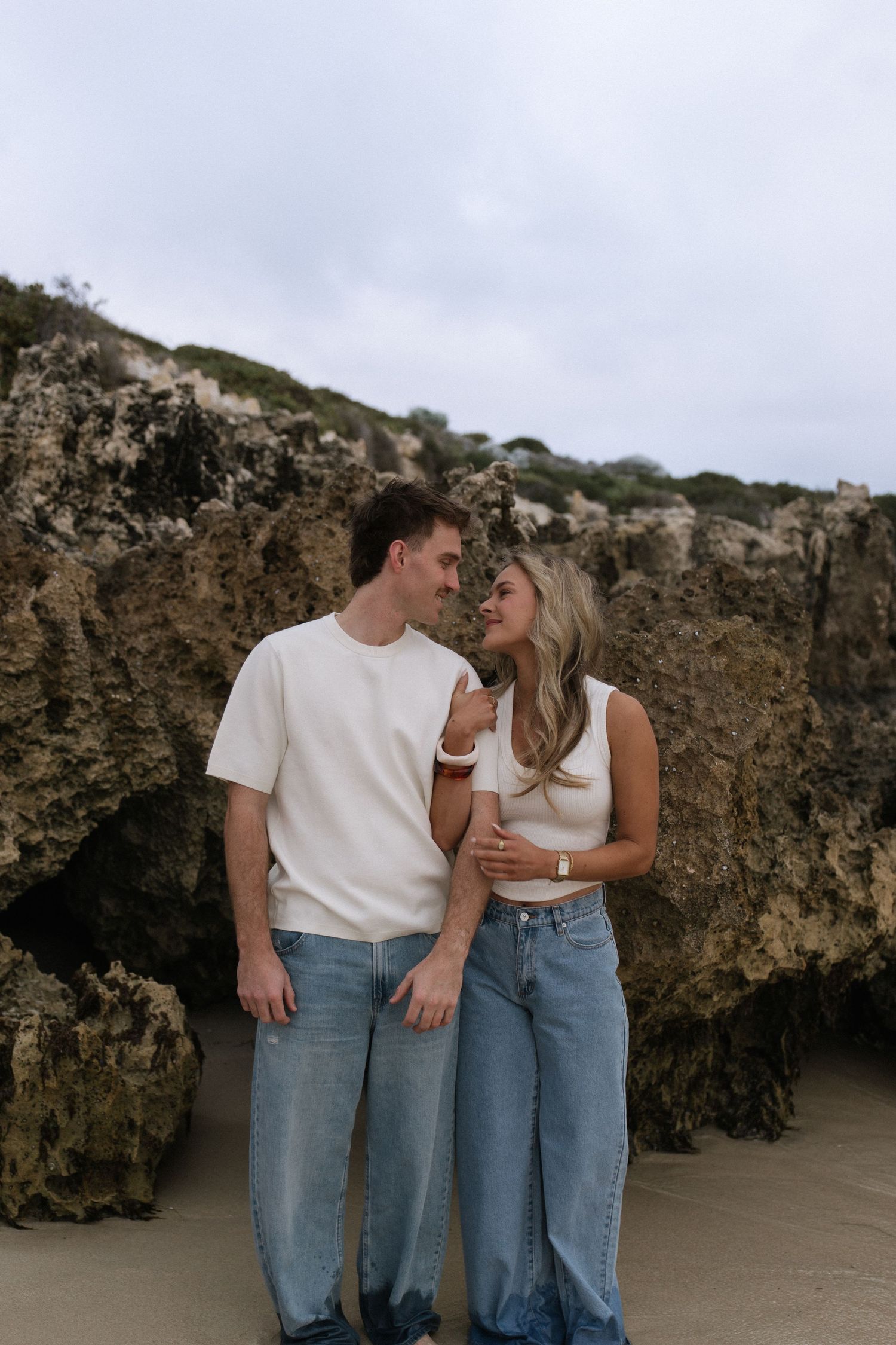 Smiling couple in white tops and jeans gazing at each other romantically against rocky beach cliffs.