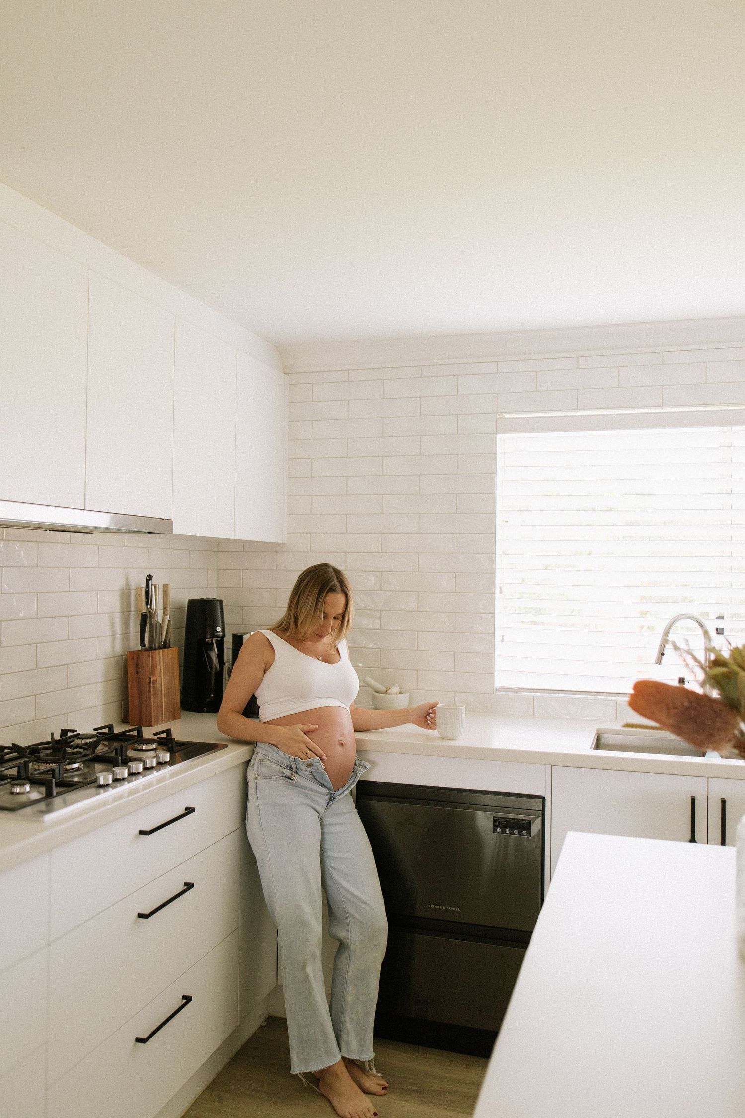 Pregnant woman in a white tank top and jeans standing in a modern white kitchen holding a mug.