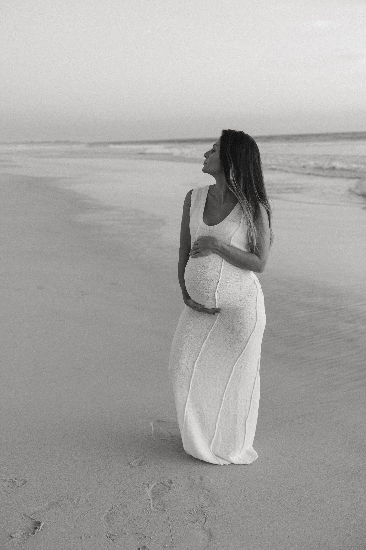 Pregnant woman in white maxi dress standing on beach at dusk, looking sideways, black and white.