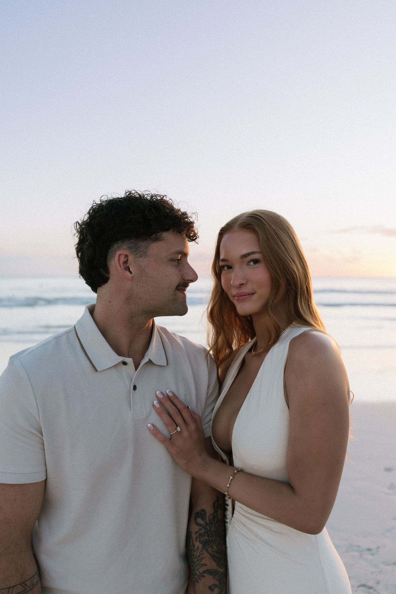 Engaged couple posing at beach sunset, woman in white dress showing ring, man in polo shirt.