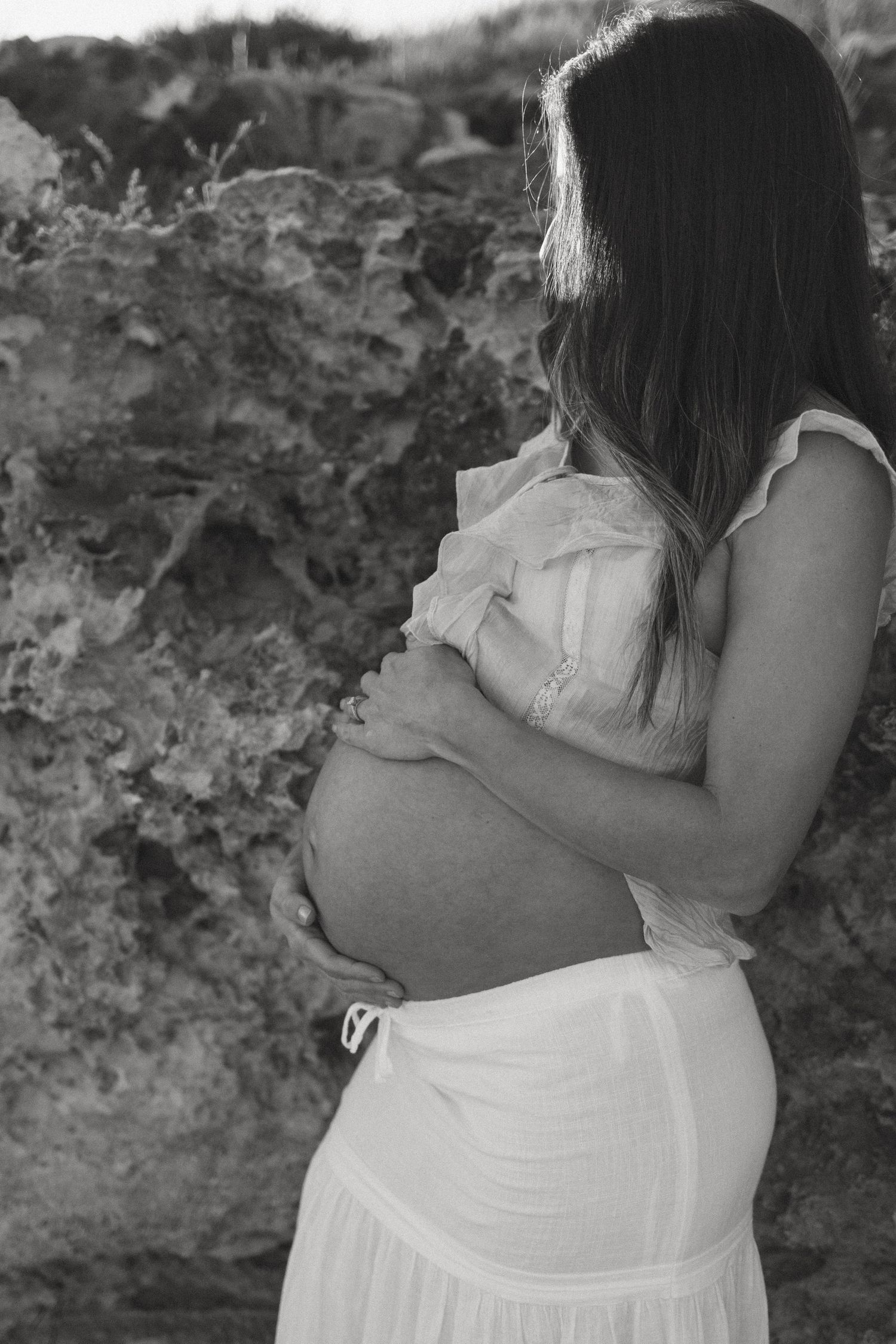 Black and white photo of pregnant woman in white dress holding belly near rocky cliff.