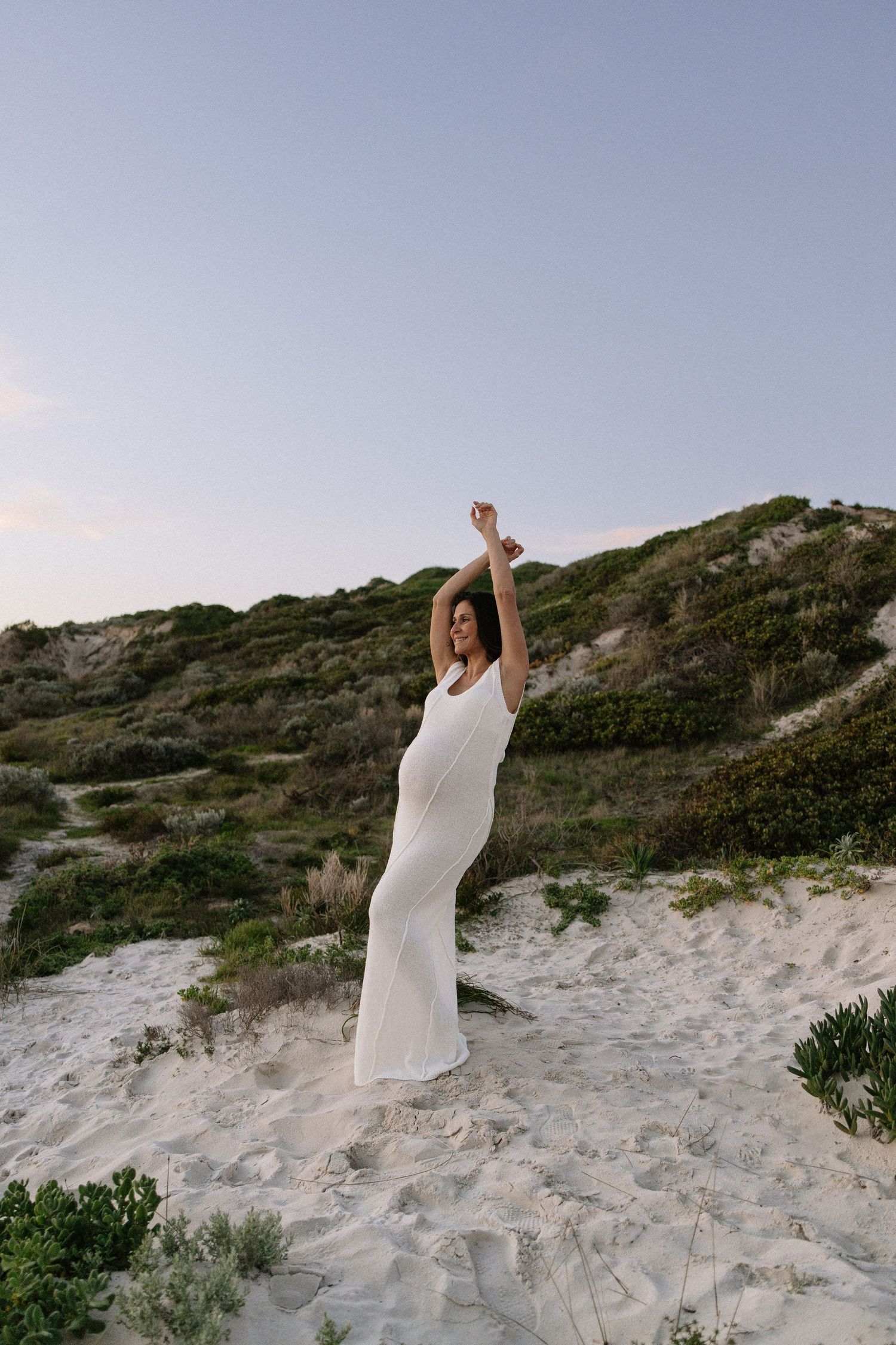 Pregnant woman in white dress raising arms on sandy beach dunes at dusk.