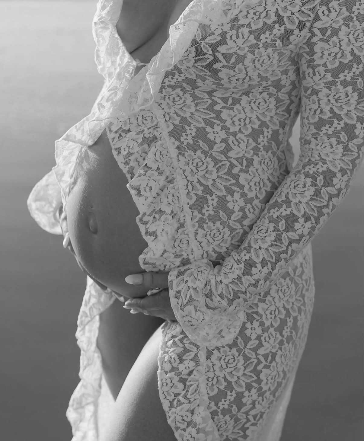 Black and white maternity photo of pregnant woman in white lace dress cradling her bare baby bump.