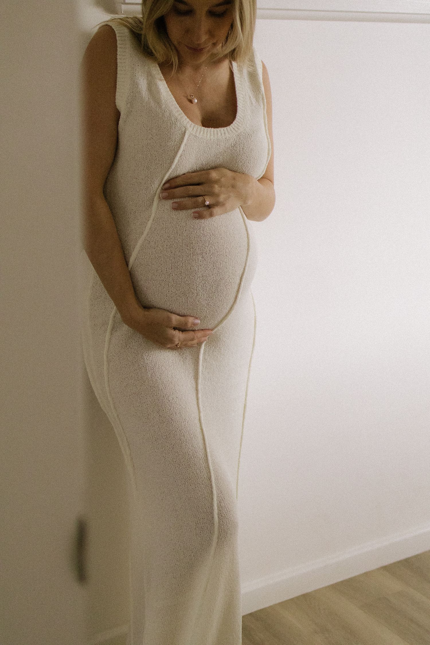 Pregnant woman in cream knit sleeveless maxi dress cradling her bump against a white wall.