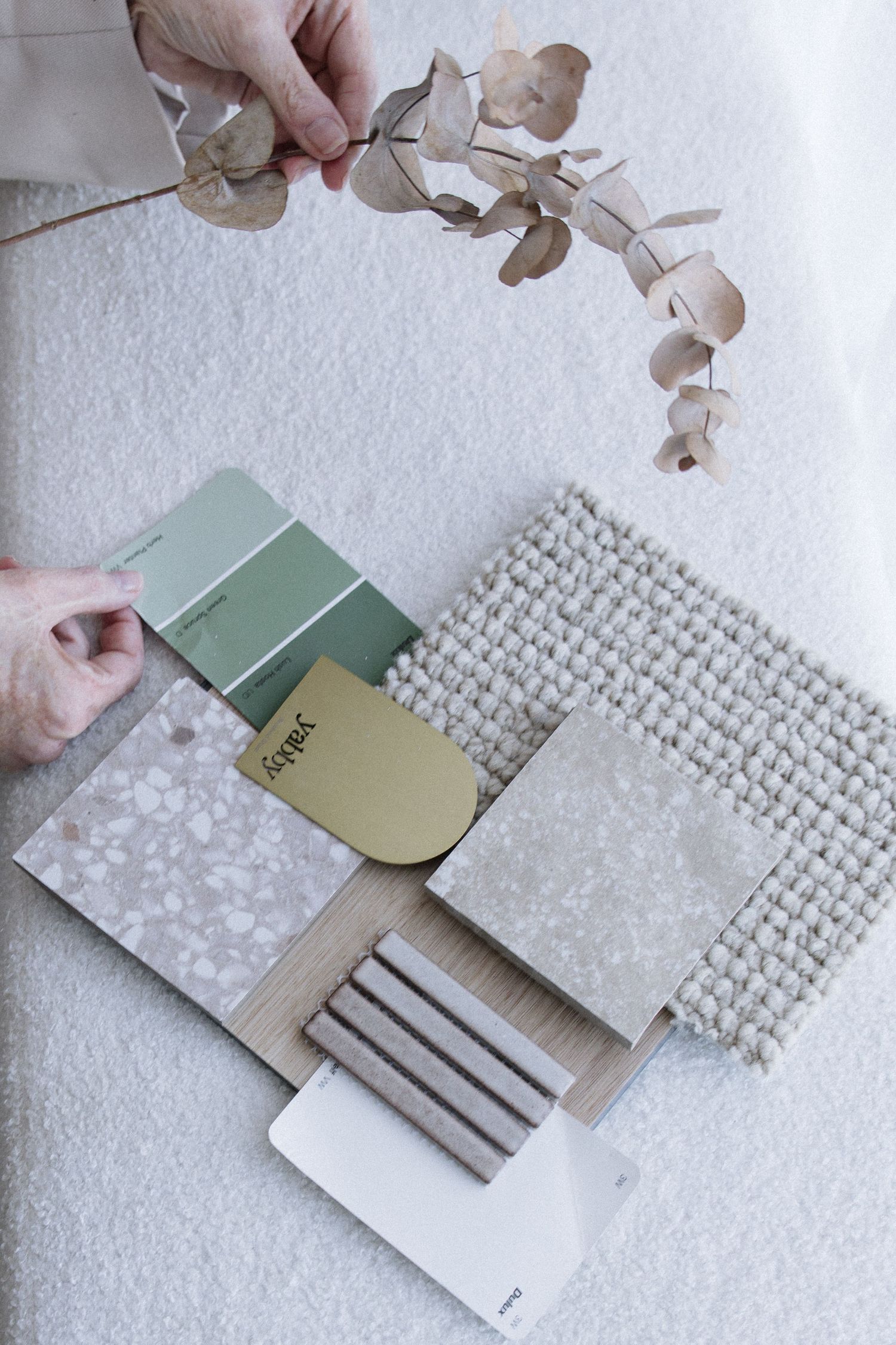 Hands arranging green paint swatches and neutral material samples with dried eucalyptus on white carpet.