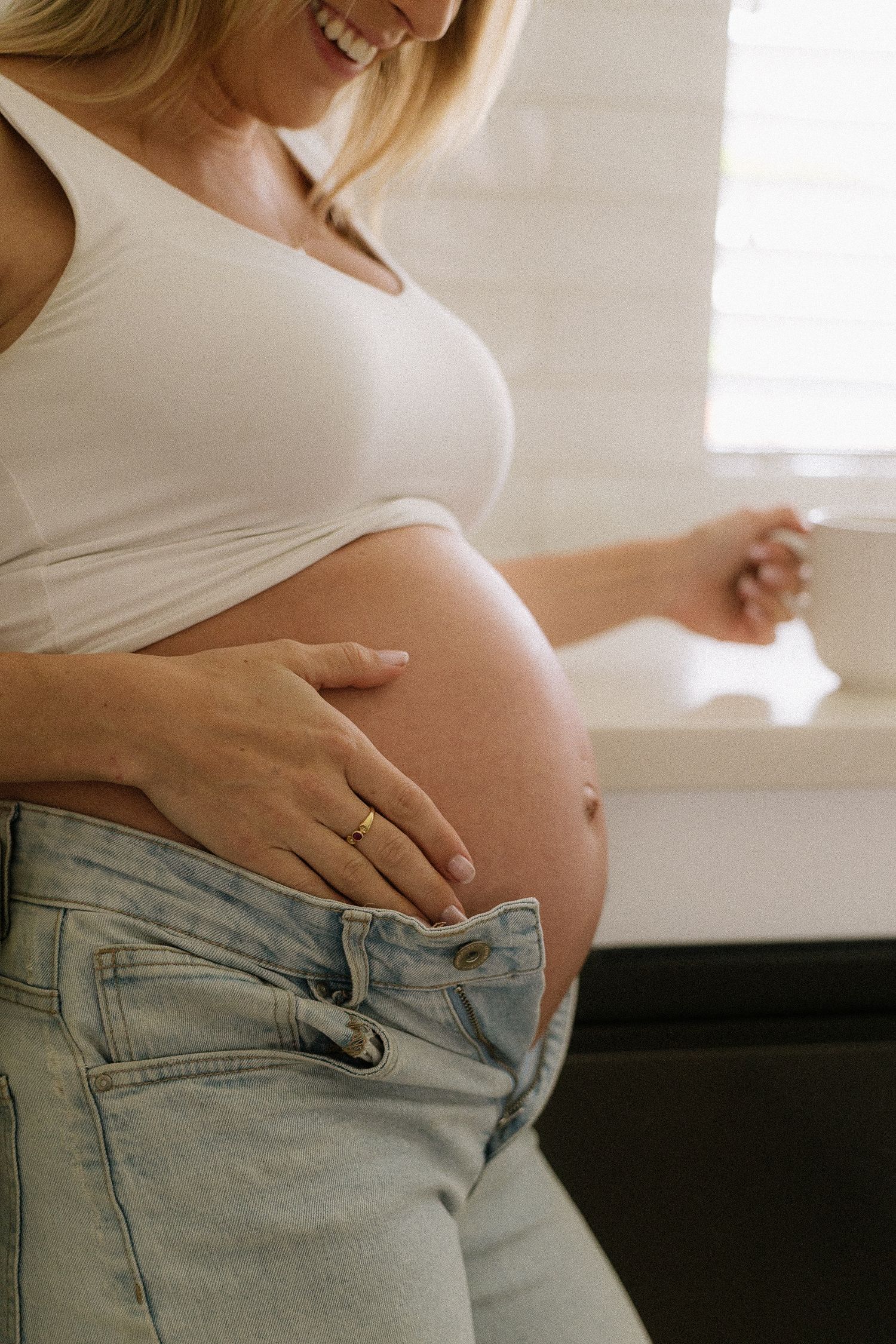 Smiling pregnant woman in a white crop top and jeans cradling her baby bump near a bright window.