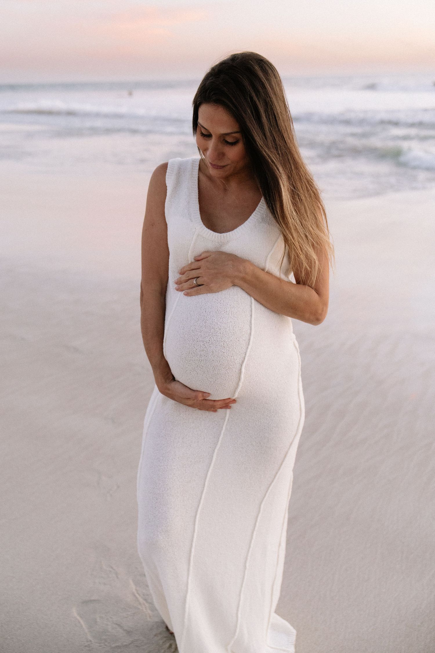 Pregnant woman in white sleeveless dress holding belly on beach at pink sunset.