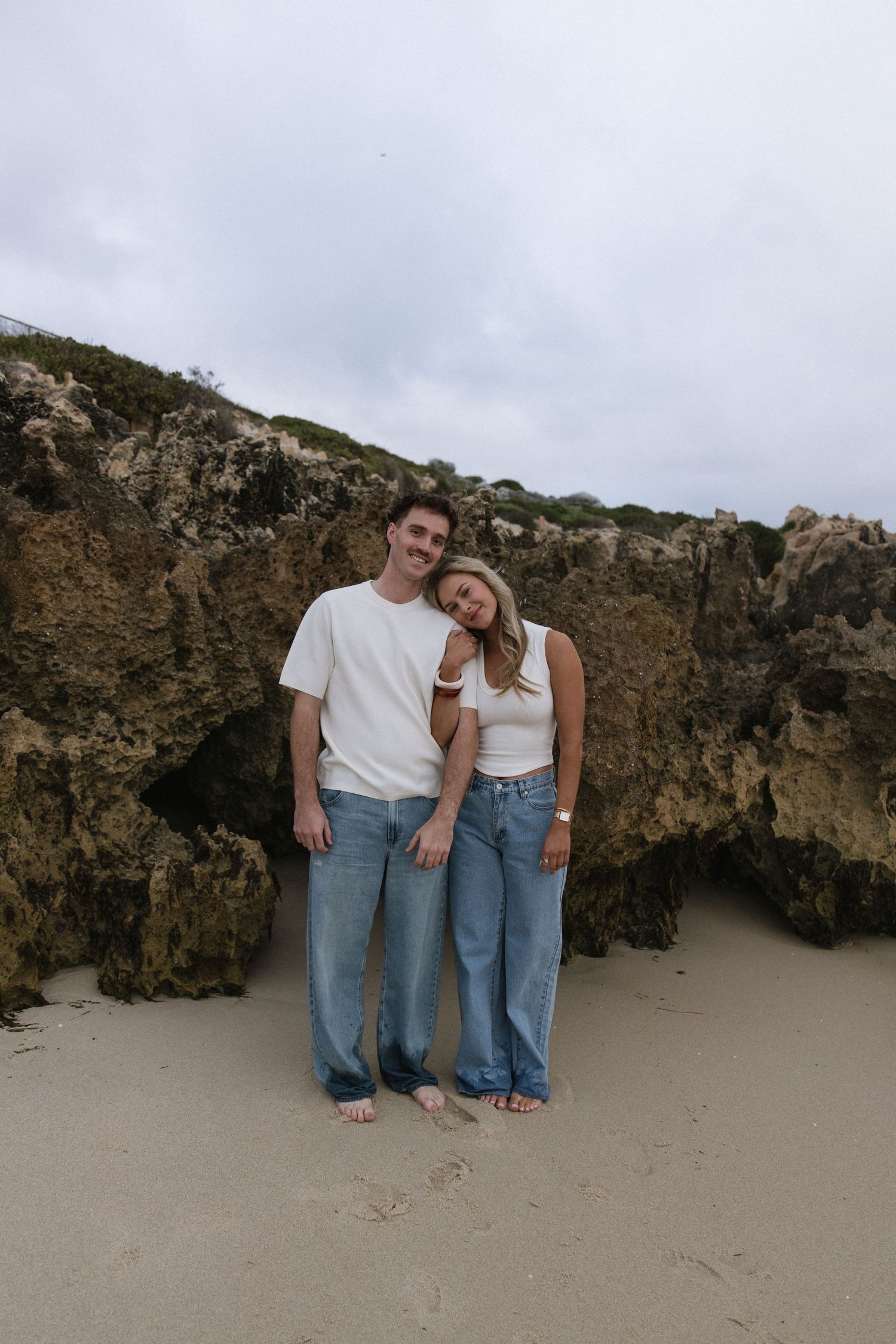 Happy couple in matching white tops and blue jeans posing together in front of rocky cliffs on a sandy beach.