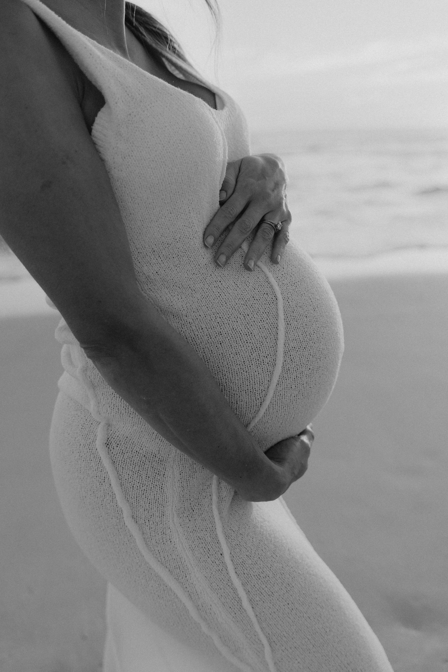 Black and white close-up of pregnant woman in white dress gently holding baby bump on beach.