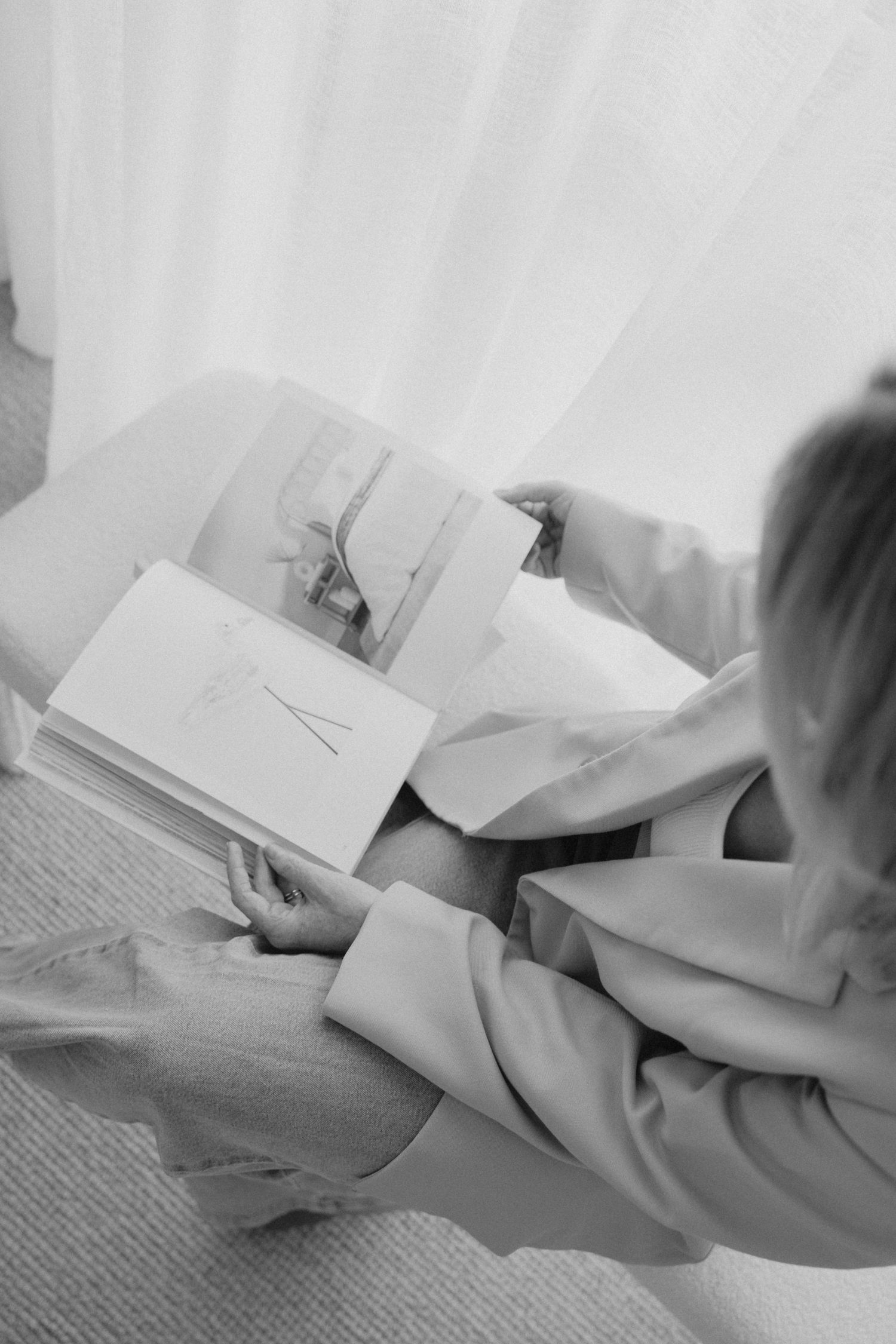 Overhead black and white shot of woman in blazer flipping through illustrated book pages near window.