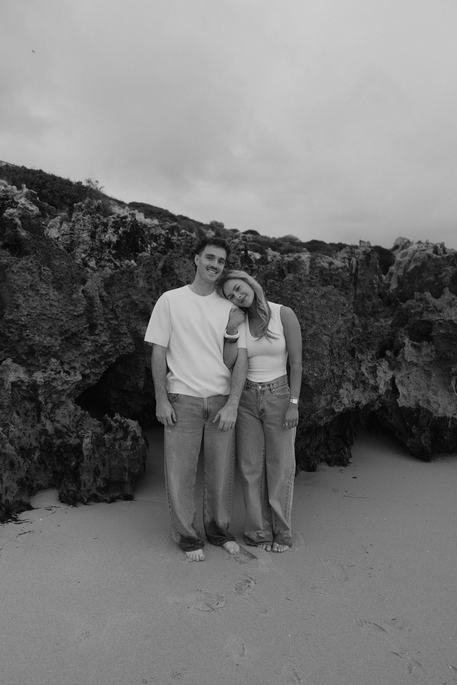 Black and white photo of a couple standing together on a sandy beach with rocky cliffs behind them.