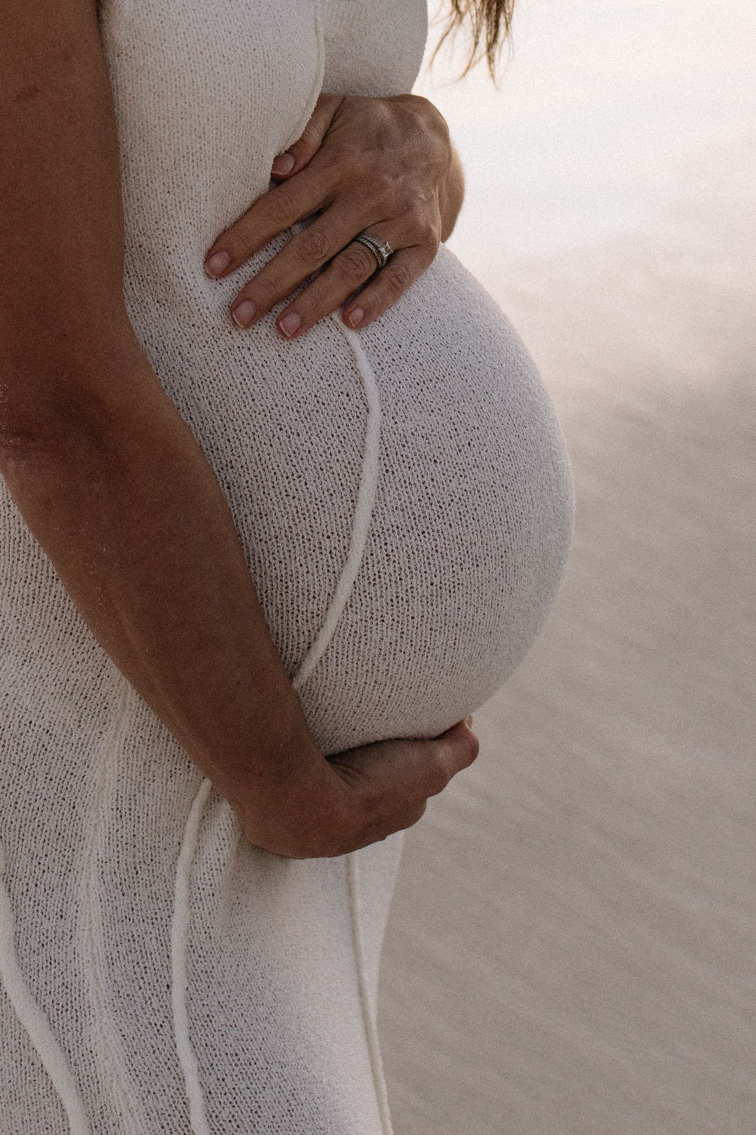 Close-up of pregnant belly in white knit dress with hands cradling bump on beach.