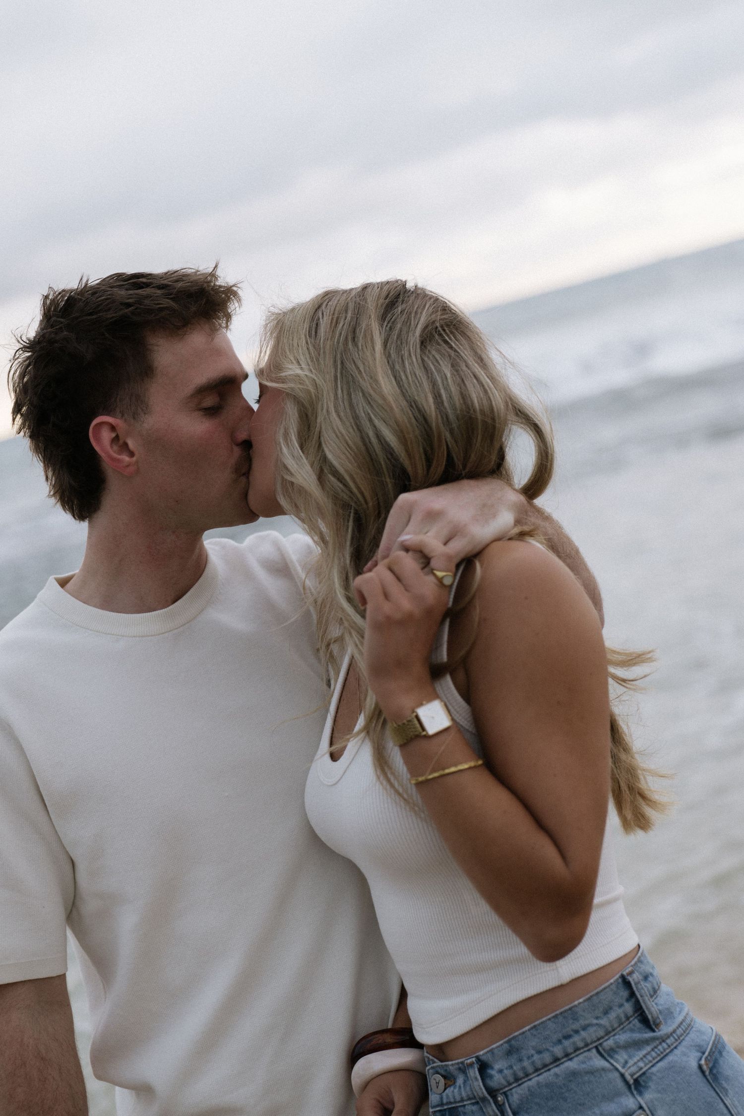 Romantic couple sharing a kiss on the beach with ocean waves behind them, woman wearing gold bracelet and watch.