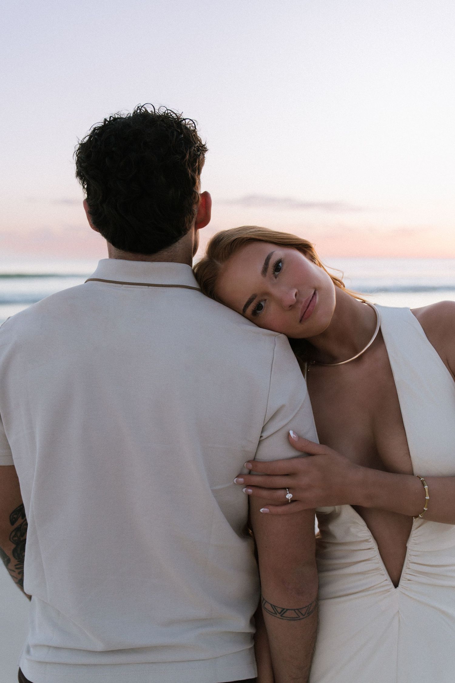 Woman in white dress leans head on man's shoulder during romantic beach engagement photo at sunset.