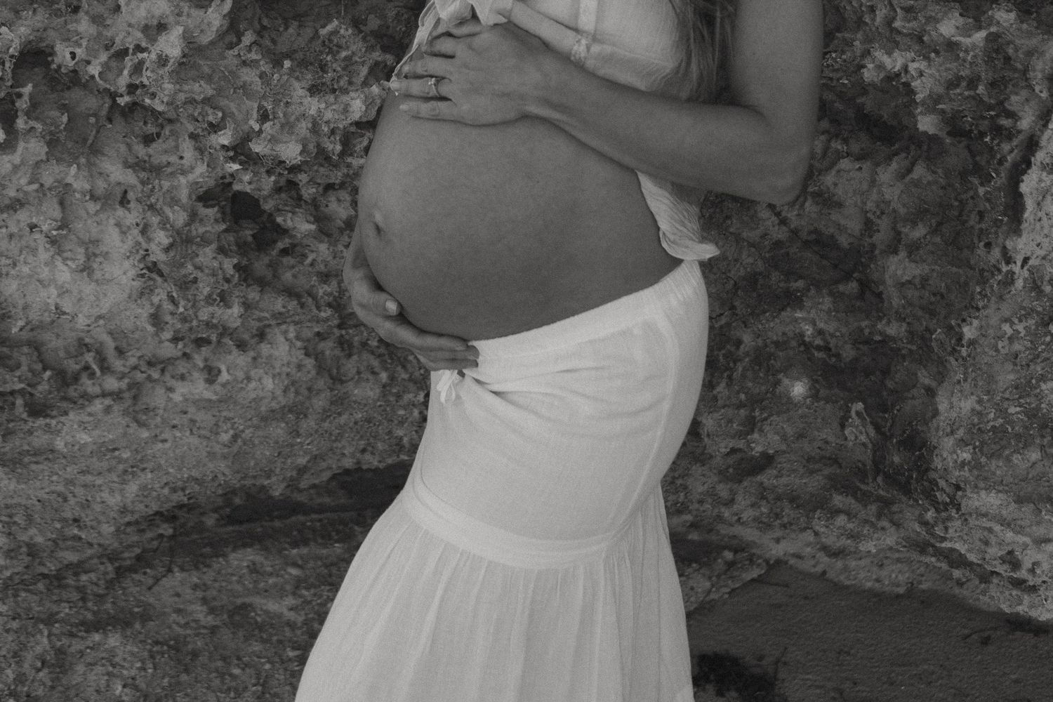 Black and white close-up of pregnant woman in white skirt holding baby bump against rocky backdrop.