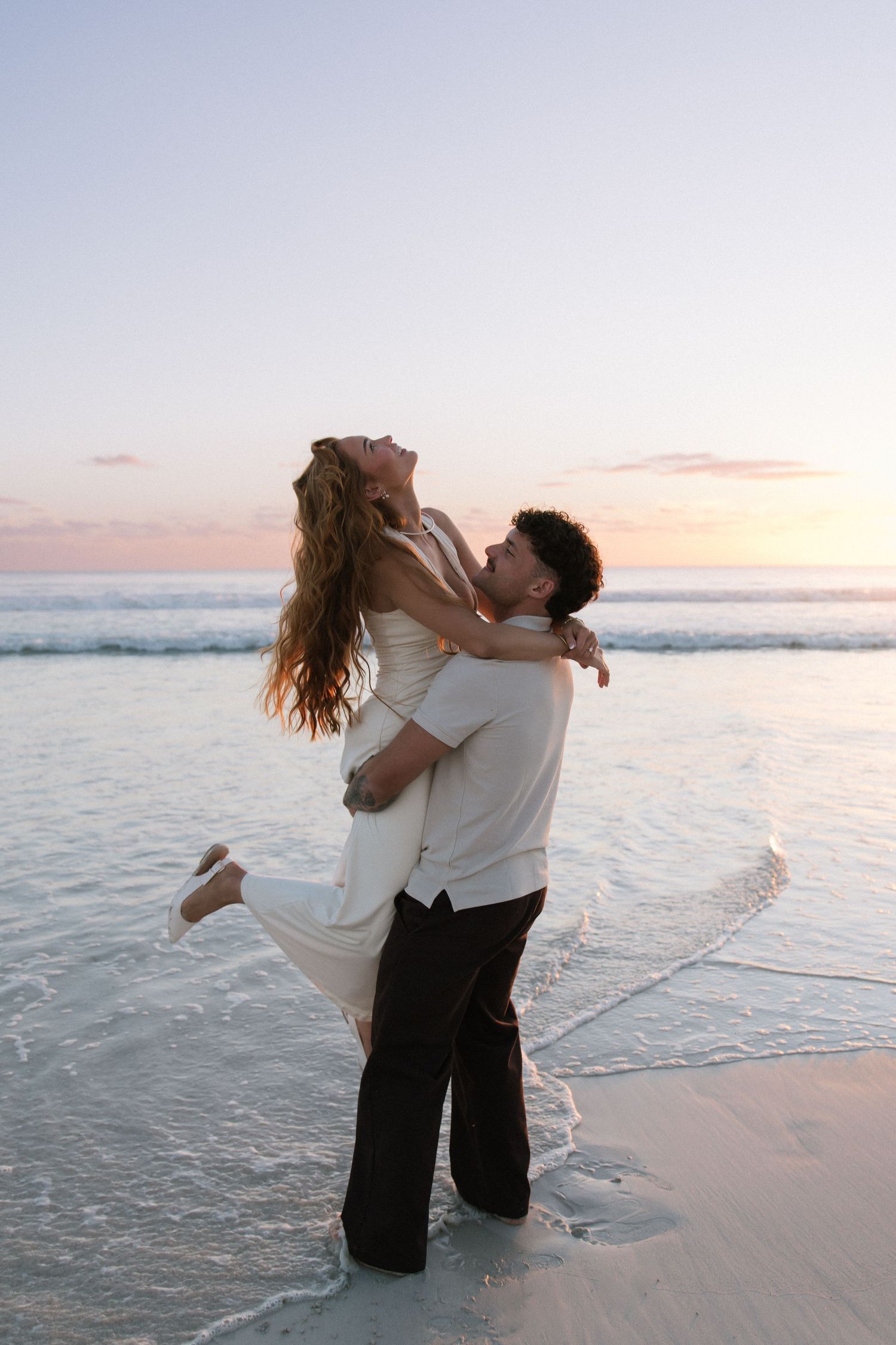 Groom lifting laughing bride in white dress on beach at colorful sunset.