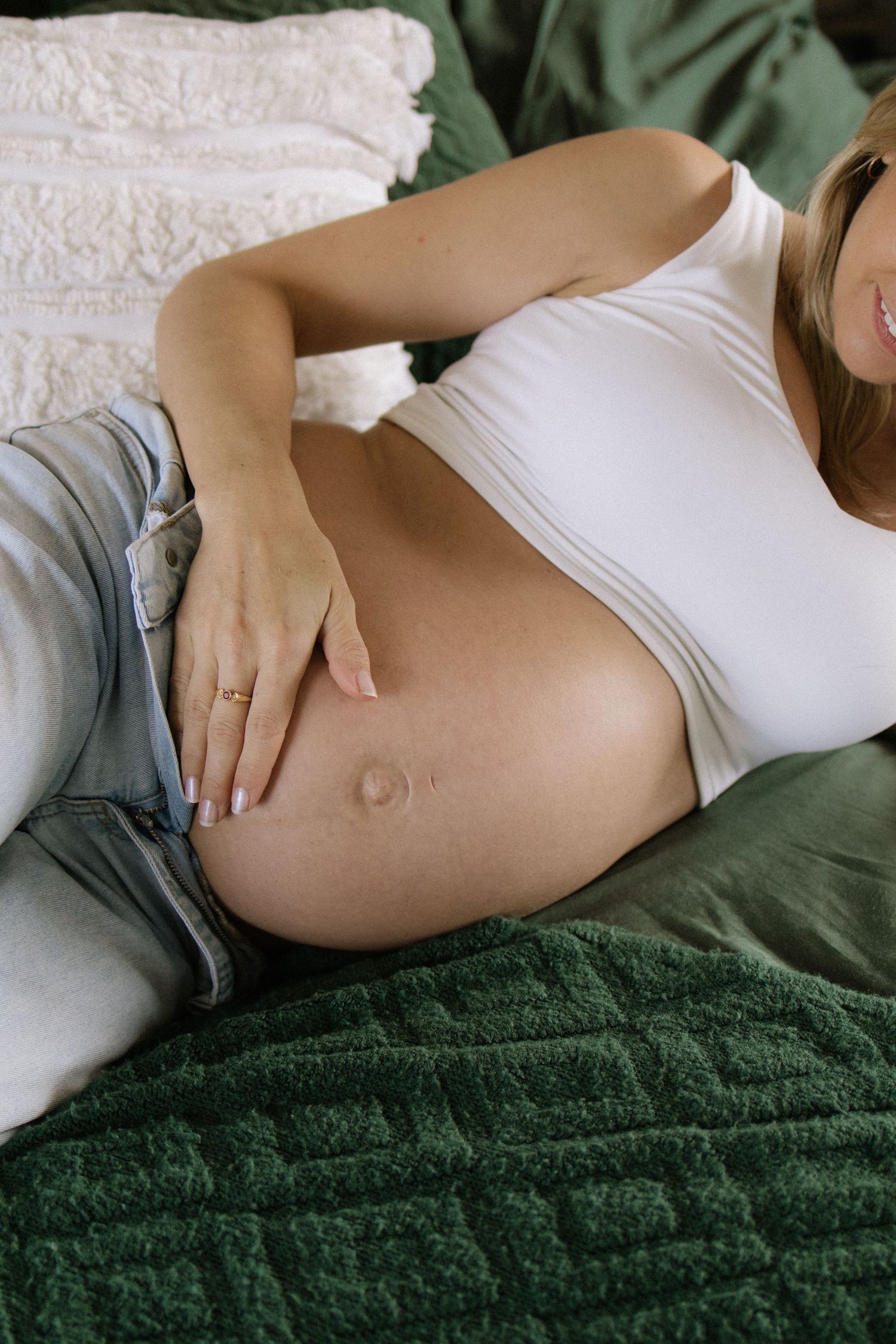 Pregnant woman in white top reclines on green bedding while gently touching her bare belly.
