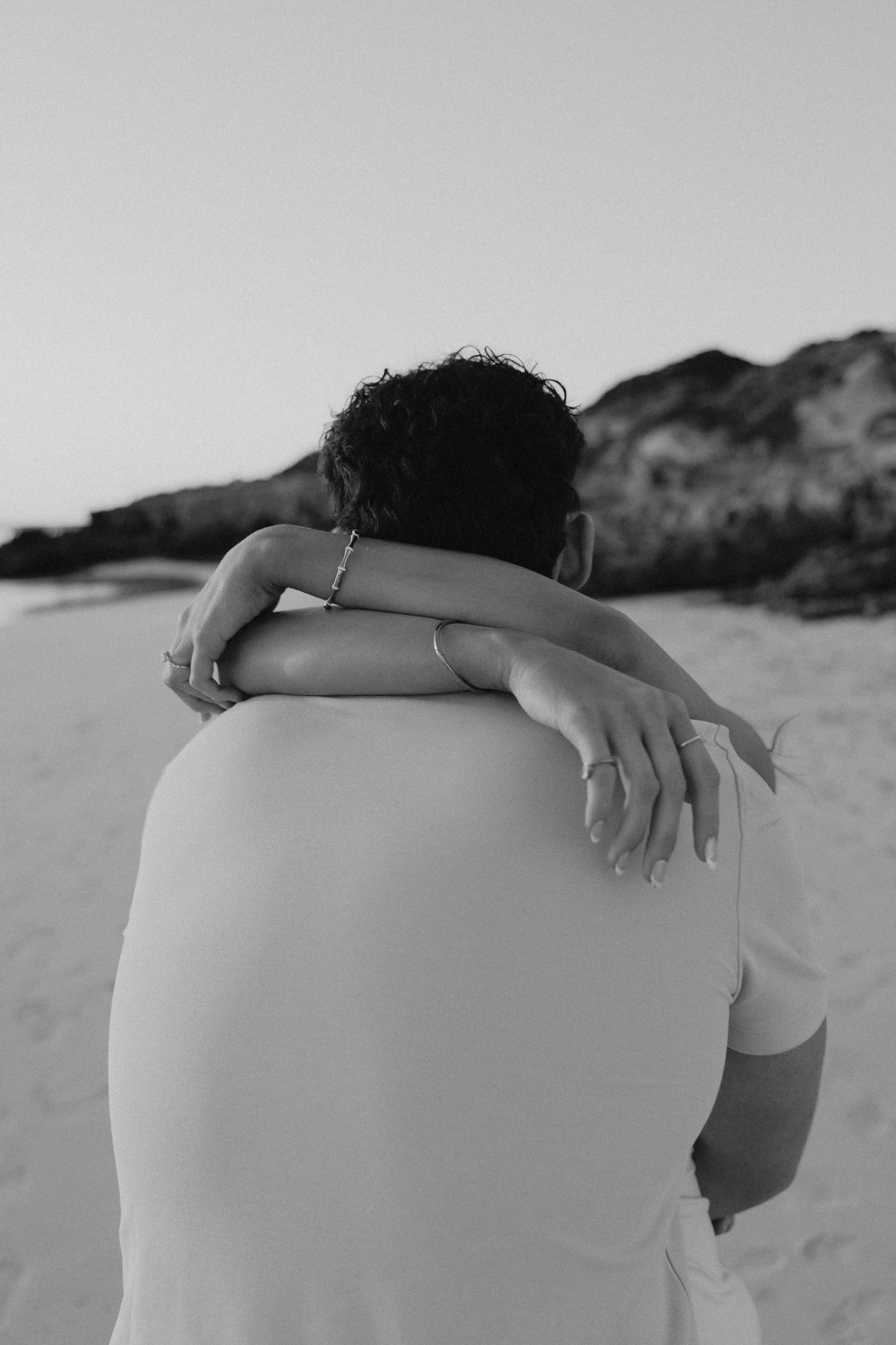 Black and white rear view of a woman in white dress embracing a man on a sandy beach.