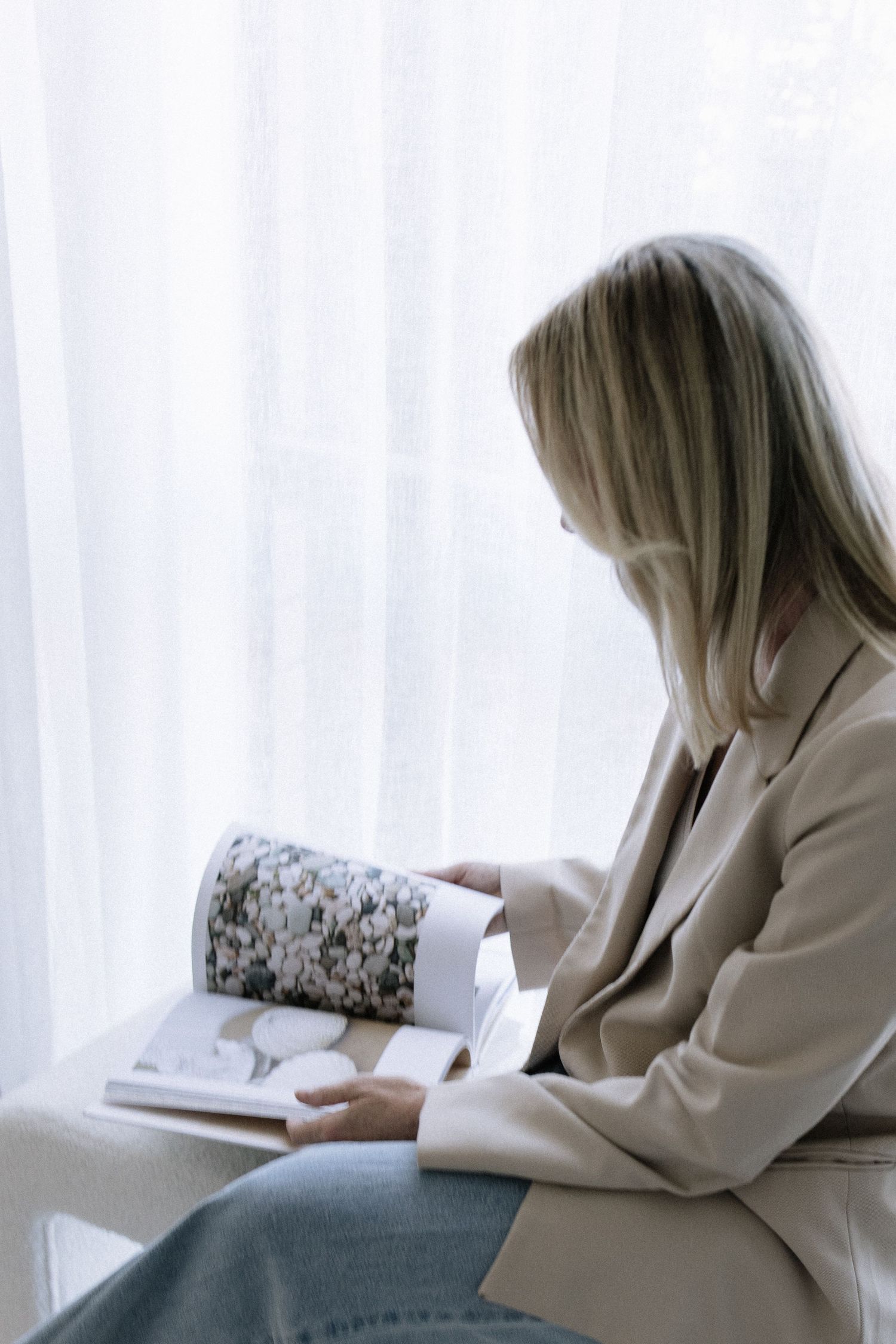 Blonde woman in beige blazer reading interior design magazine while seated near sheer white curtains.
