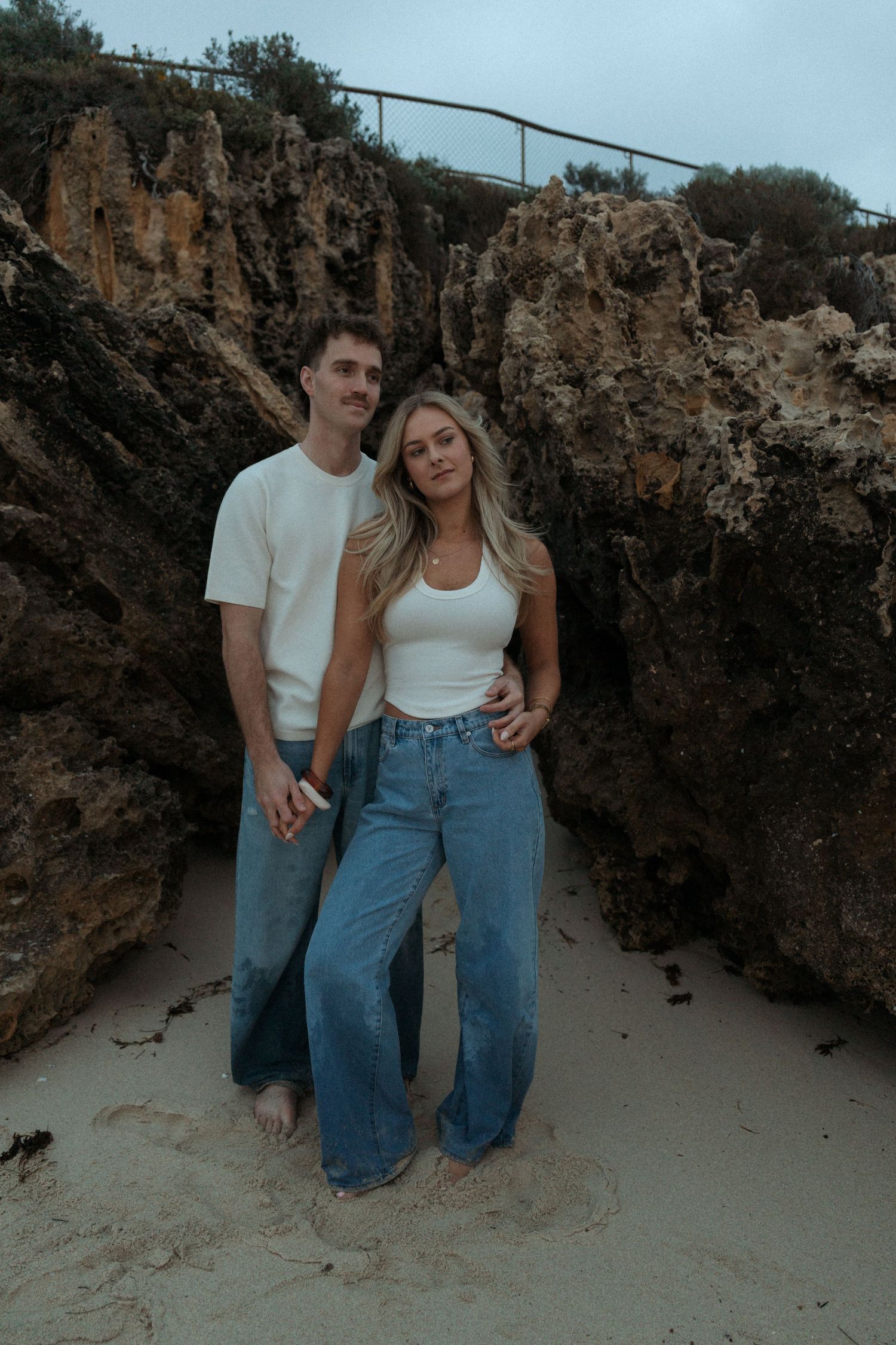 Couple posing together between rocky cliffs on a sandy beach, both wearing white tops and blue jeans.