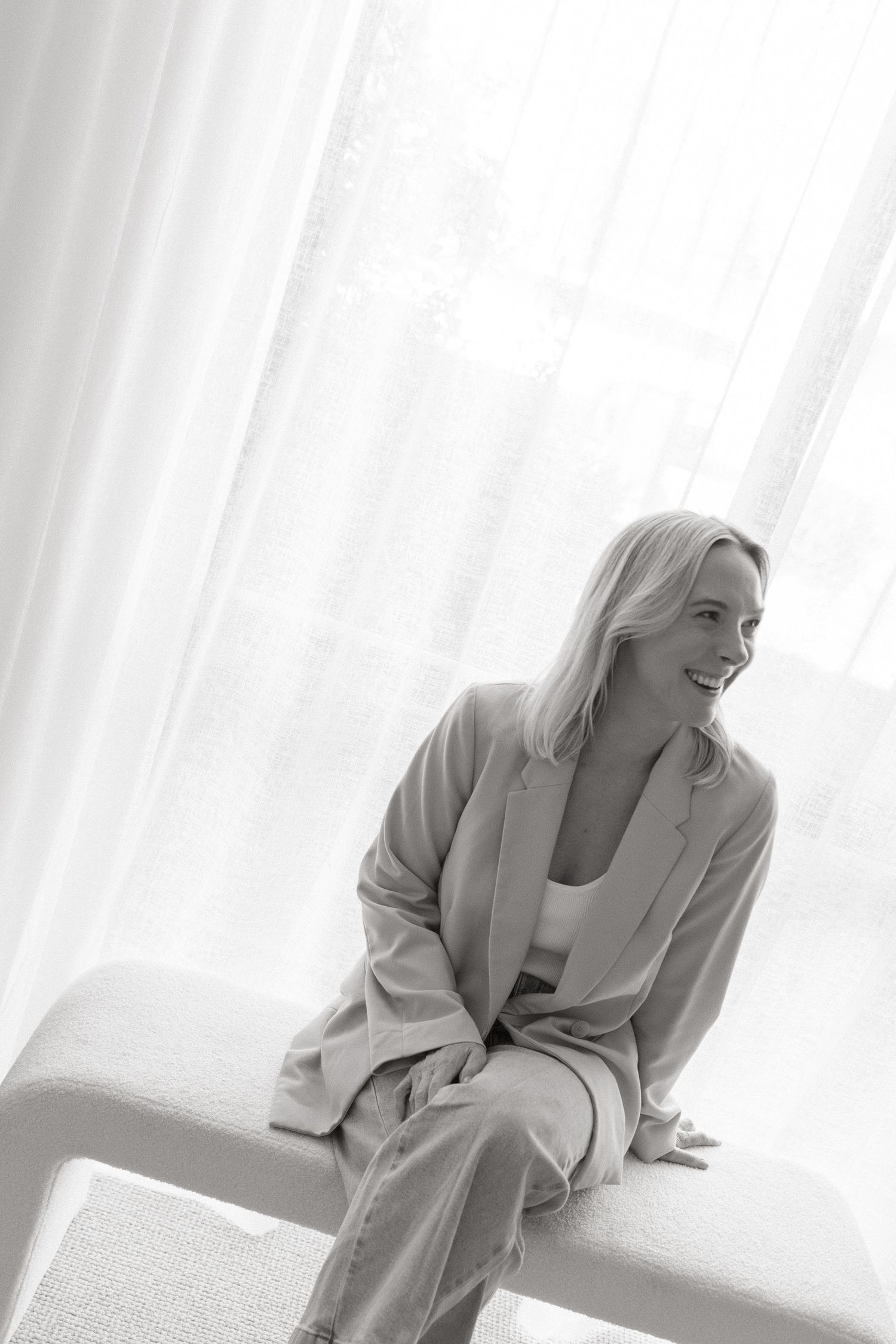 Black and white portrait of smiling blonde woman in blazer sitting on white bench near sheer curtains.