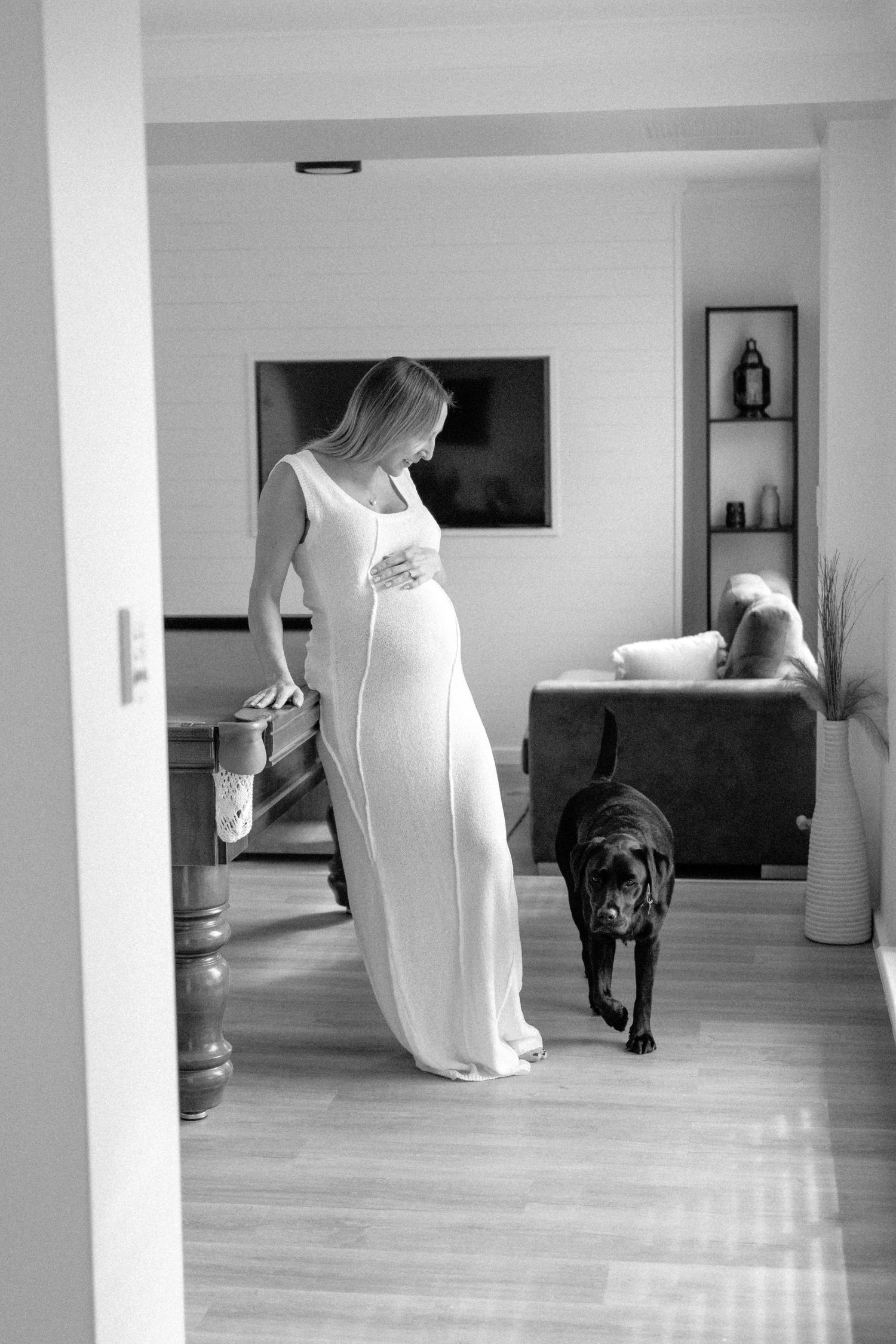 Black and white photo of pregnant woman in white maxi dress walking with black Labrador indoors.